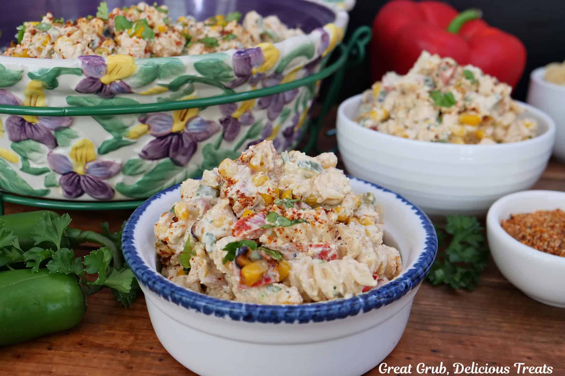A wood surface with two white bowls filled with a serving of street corn pasta salad with a floral bowl off to the side with the pasta salad in it.