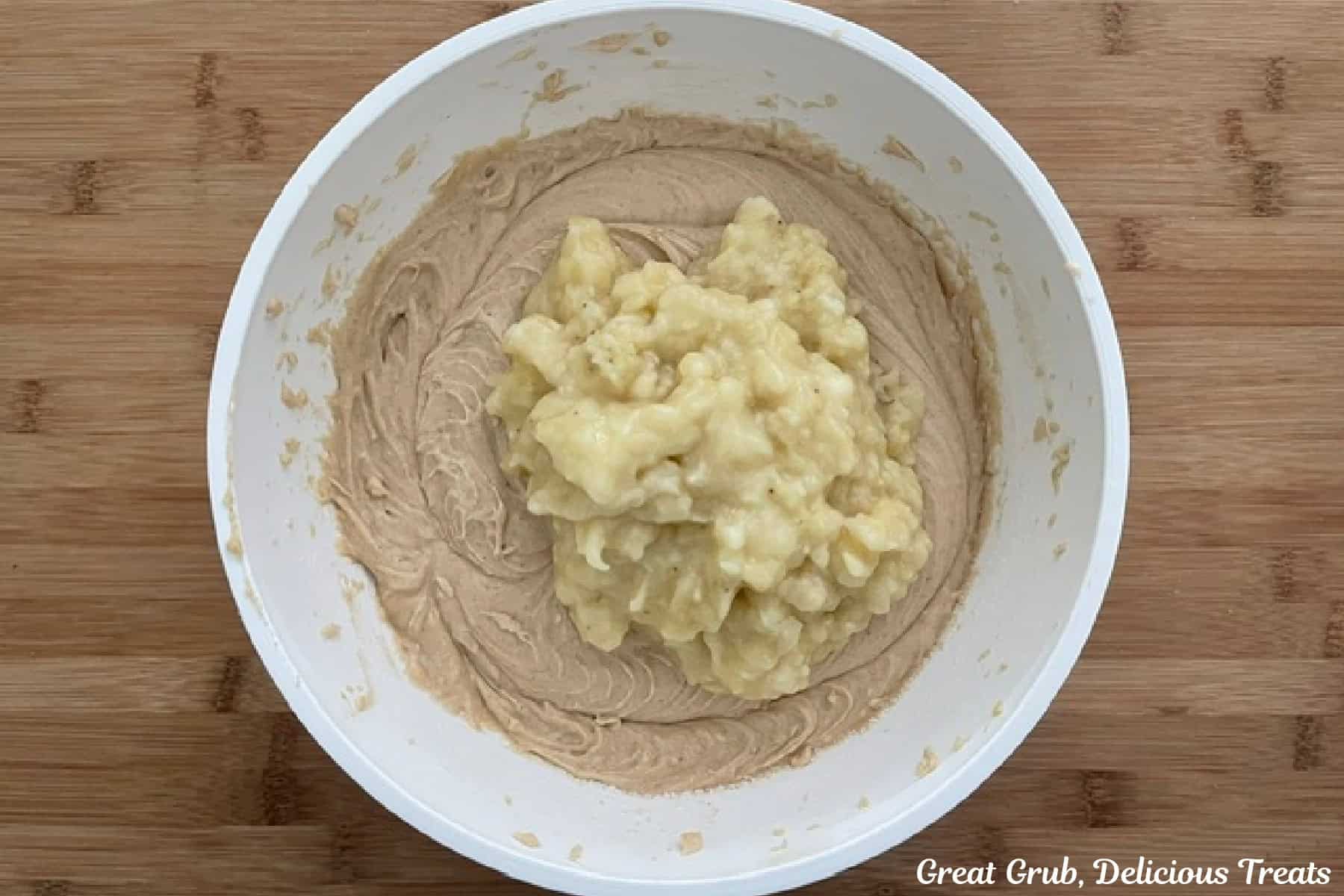 A wood surface with a mixing bowl filled with banana muffin batter with mashed bananas about to be stirred into the batter.