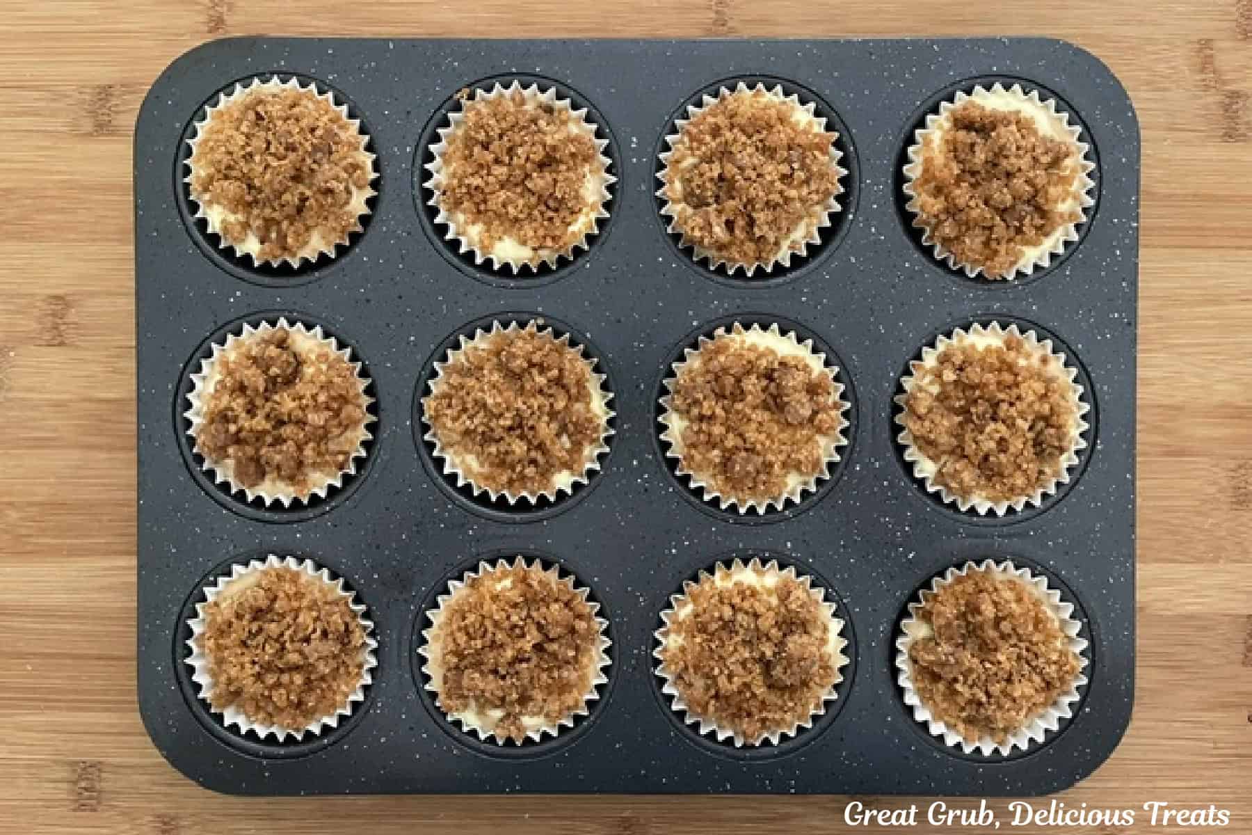 A wood surface with a 12-count muffin tin filled with banana cream cheese muffin batter and topped with streusel topping.
