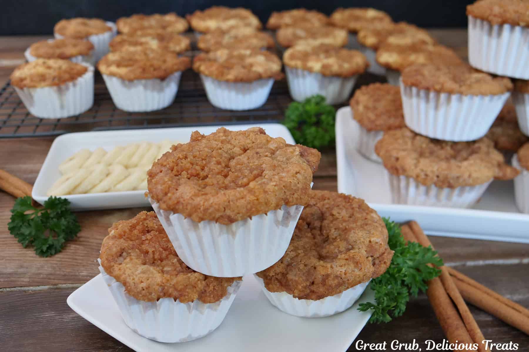 A wood surface with a dozen and a half banana cream cheese muffins on it with sliced bananas on a white plate and a few cinnamon sticks placed on the side.