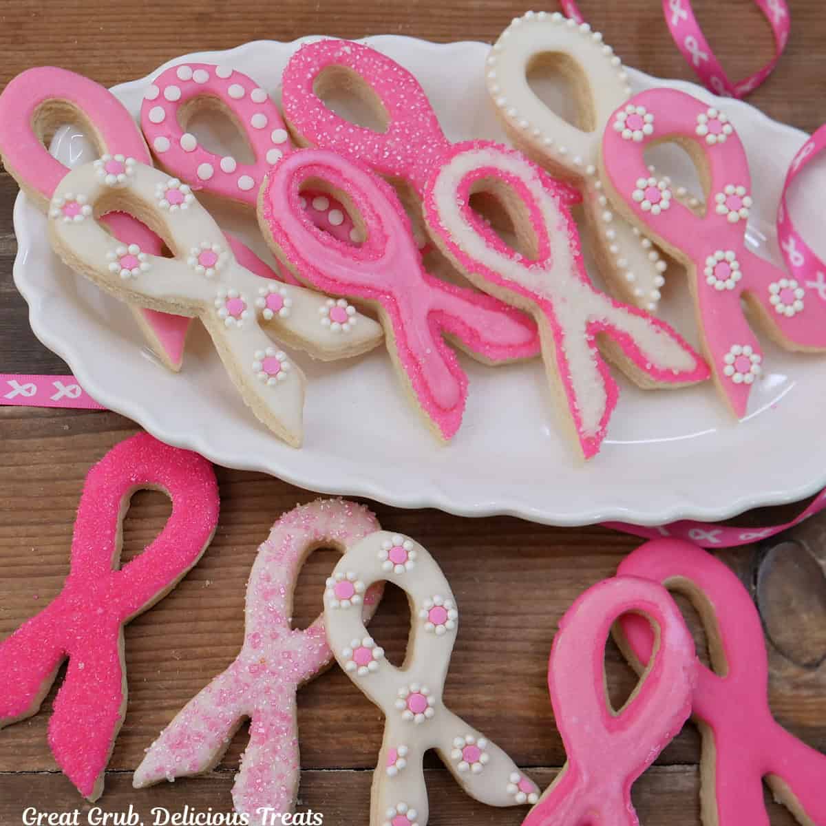 Breast Cancer Awareness Ribbon sugar cookies on a white oval plate and some placed on a wood surface featuring white and pink decorated cookies.