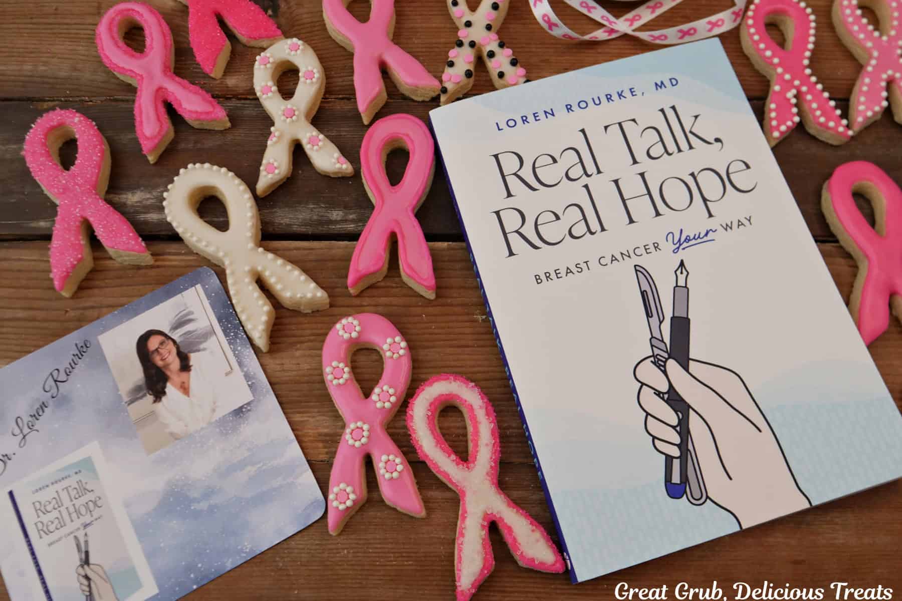 A wood surface with Dr. Loren Rourke's book, "Real Talk, Real Hope" and her photo on a card surrounded by breast cancer awareness ribbon sugar cookies.
