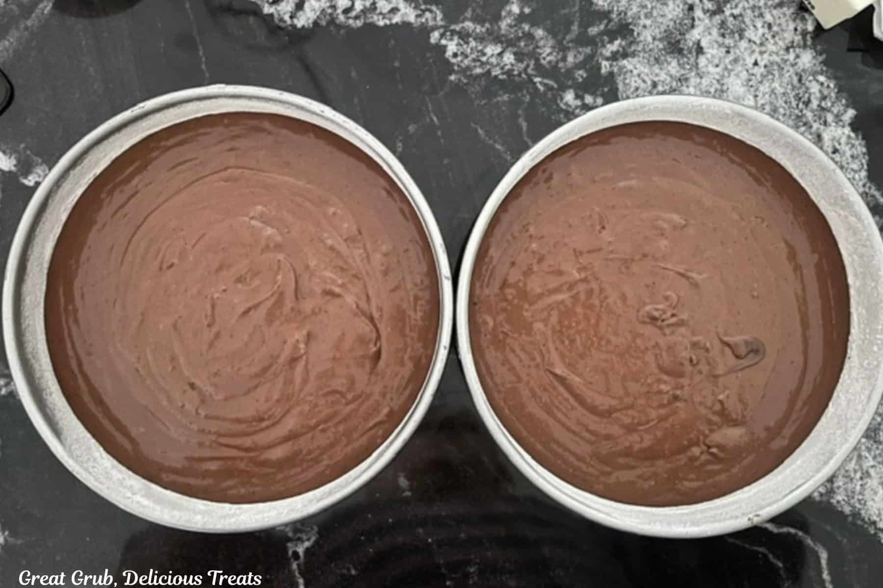 A black granite surface with two silver round cake pans filled with chocolate cake batter.