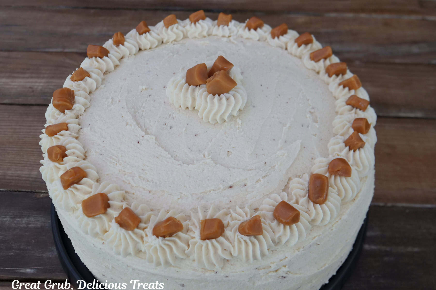 A wood surface with a salted caramel frosted two layer cake with pieces of soft caramel around the top on the frosting.