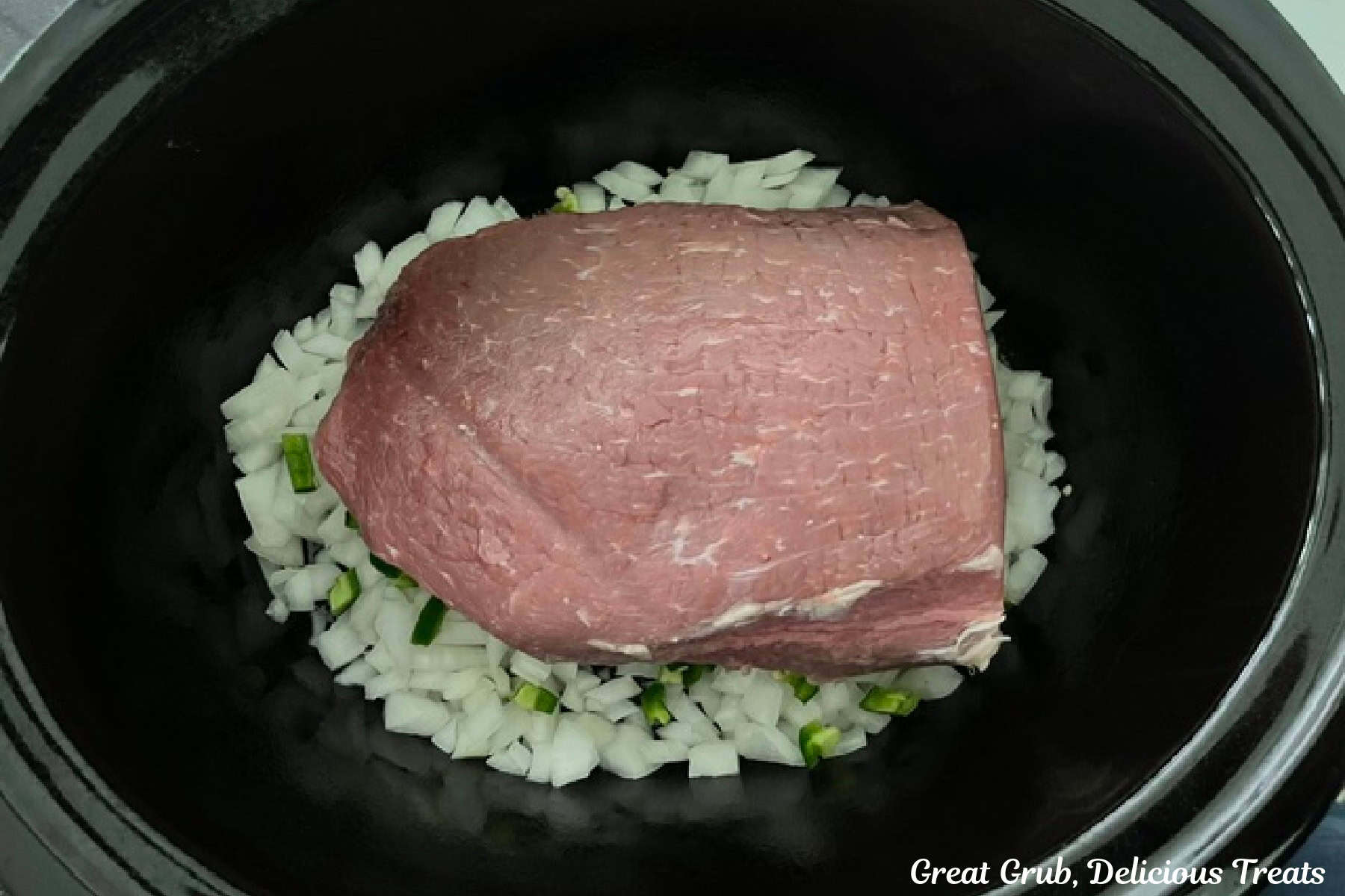 A black crock pot with diced onions and jalapenos, and a whole round roast placed in it.