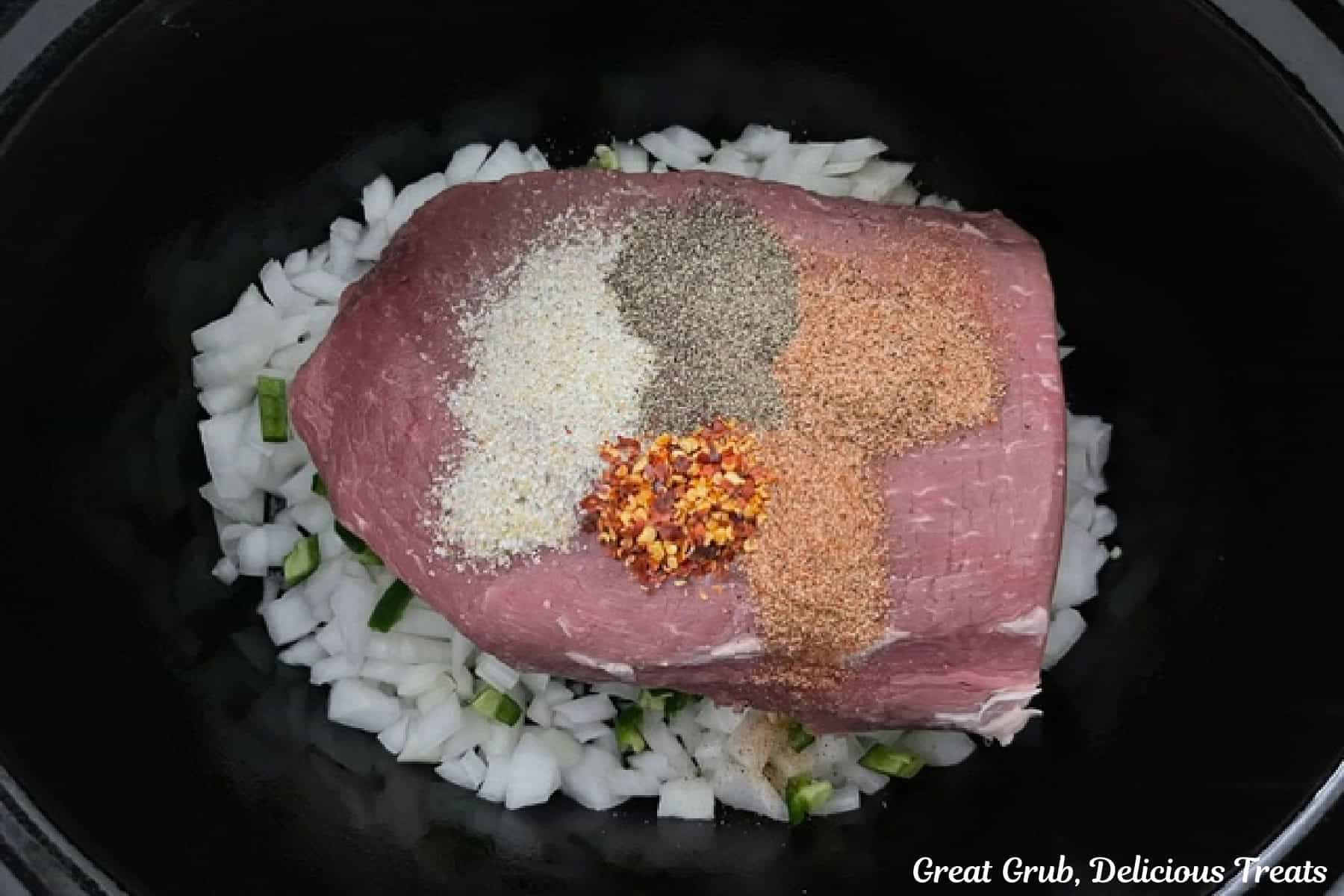 A black crockpot with diced onions, jalapenos, a round roast, and seasonings on top of the roast.