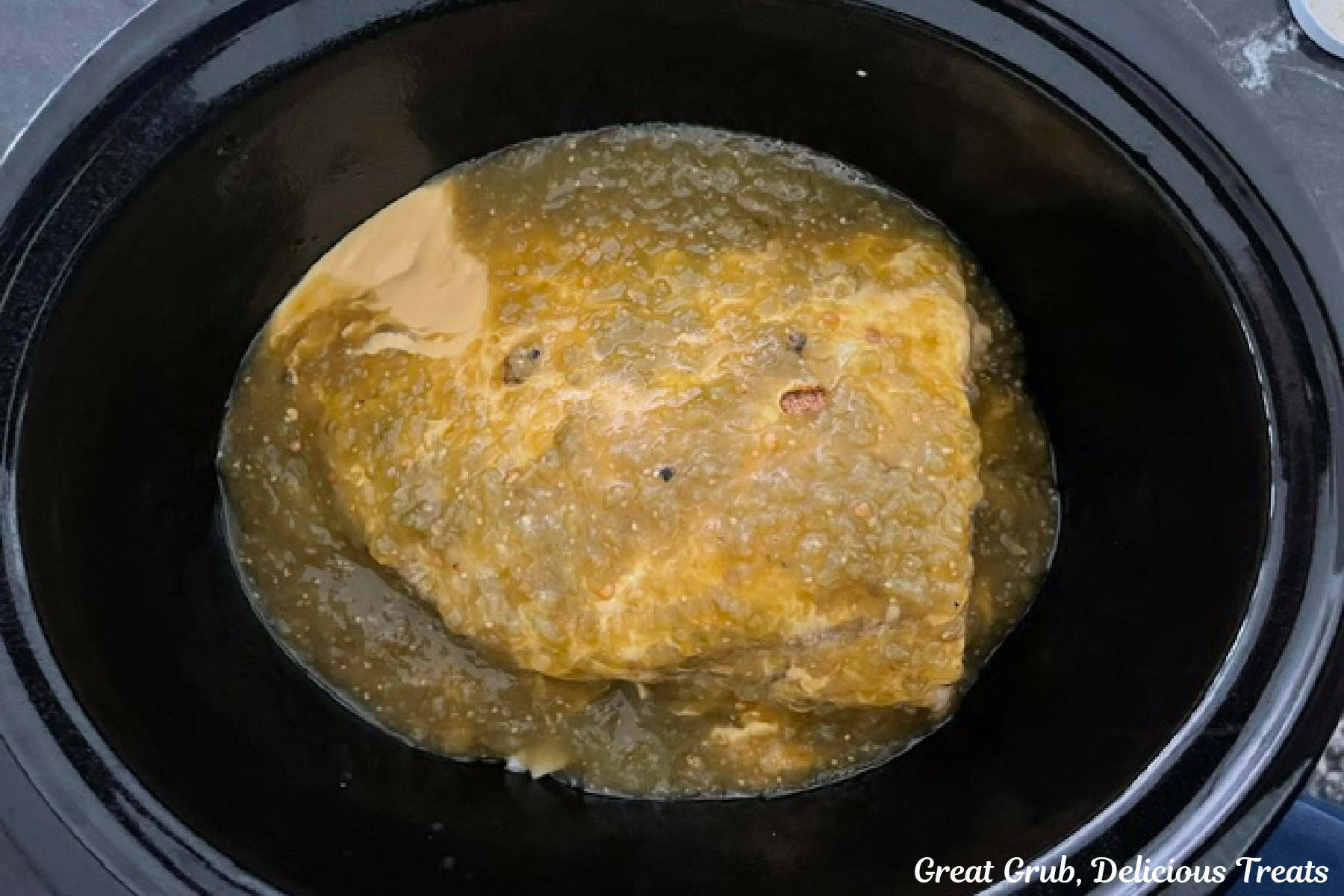 A black crockpot with round roast covered with seasonings, queso, and salsa verde before being slow cooked.