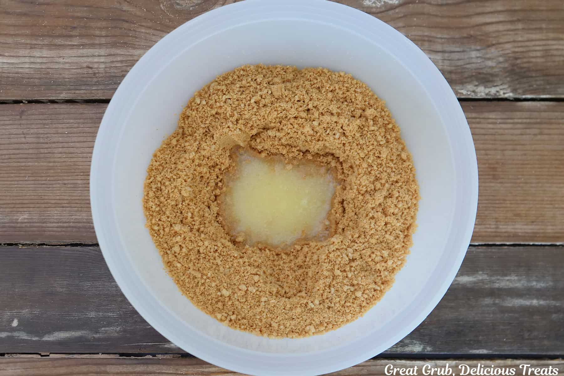 A wood surface with a white mixing bowl filled with crushed peanut butter cookies and melted butter in it.