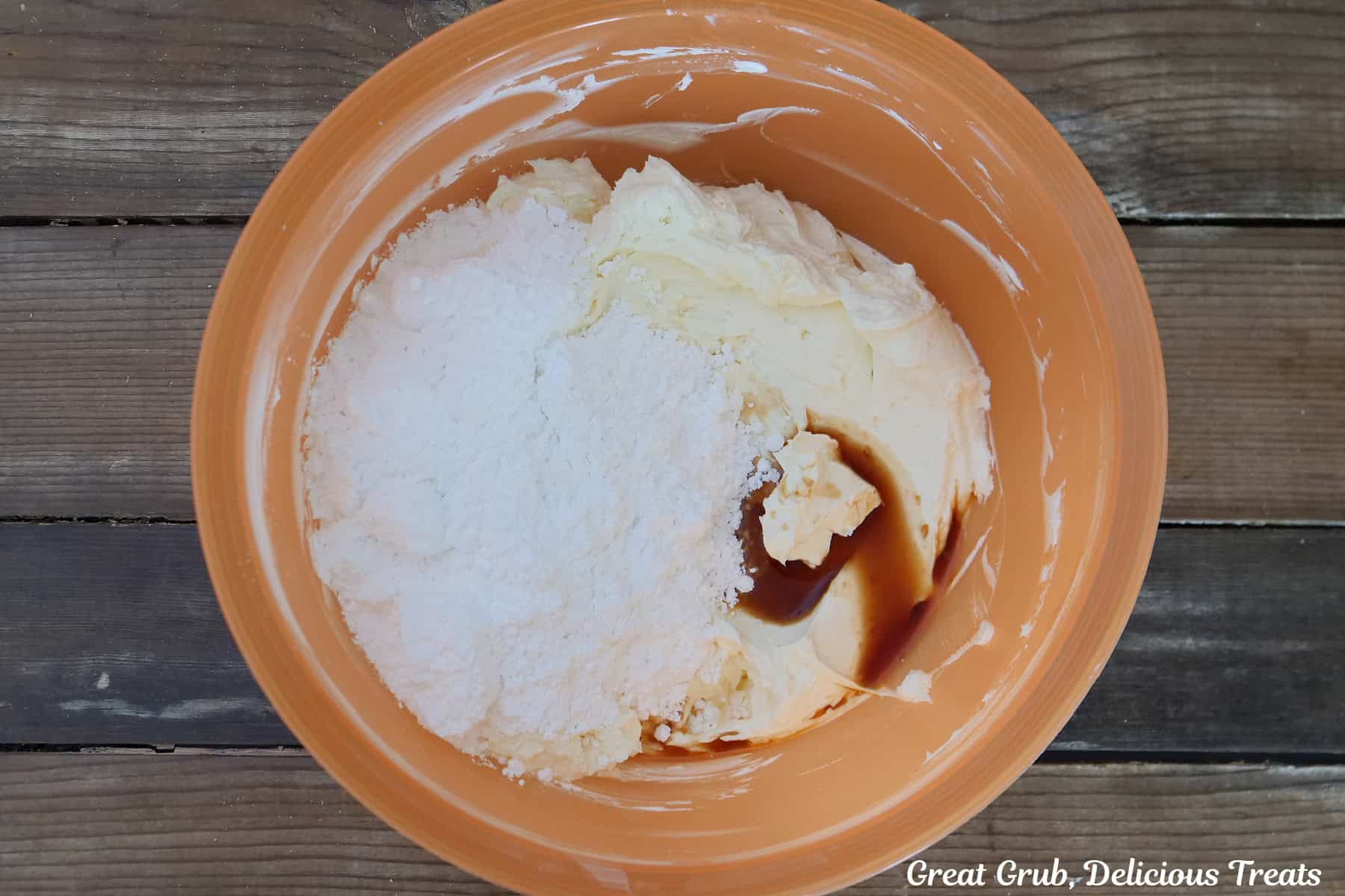 A wood surface with an orange mixing bowl filled with cream cheese, powdered sugar, and vanilla in it.