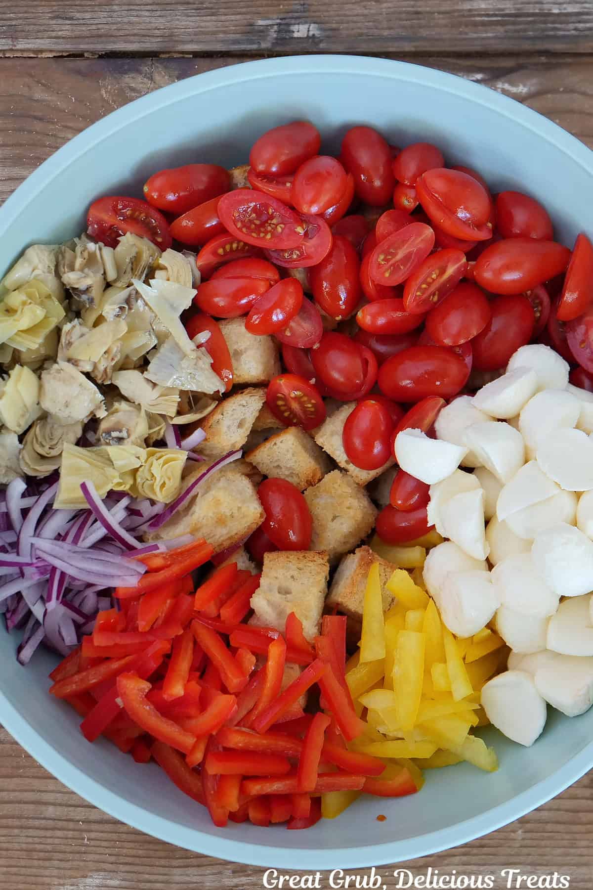 A wood surface with a light blue bowl filled with all the ingredients to make panzanella salad placed in it.