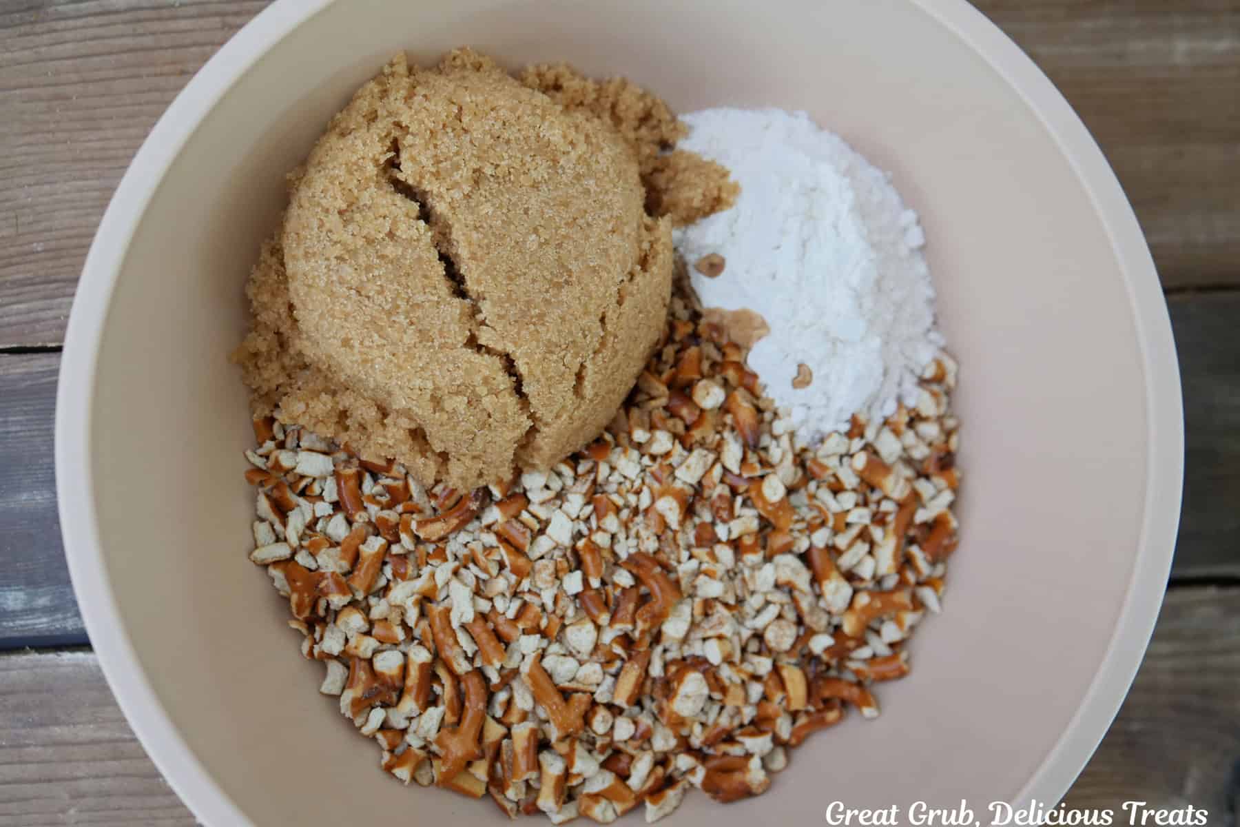 A wood surface with a white bowl filled with crushed pretzels, brown sugar, and flour, before being combined.