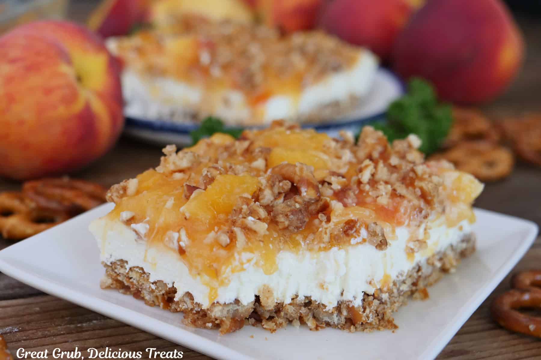 A white plate with a serving of peach pretzel dessert on it with another slice and a few peaches in the background.