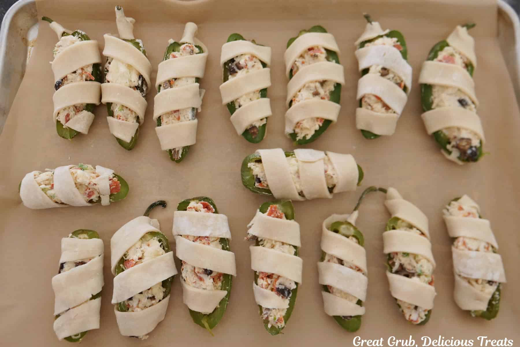 A baking sheet lined with parchment paper with sixteen pepperoni pizza jalapeno poppers wrapped in strips of pie crust before being baked.