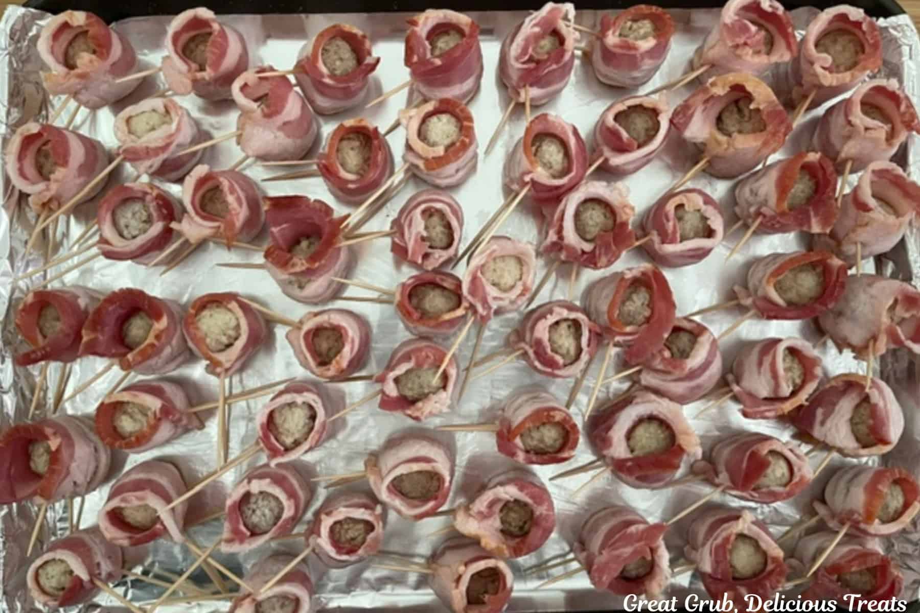 A foil lined baking sheet with about fifty bacon wrapped meatballs on it with toothpicks holding the bacon in place.