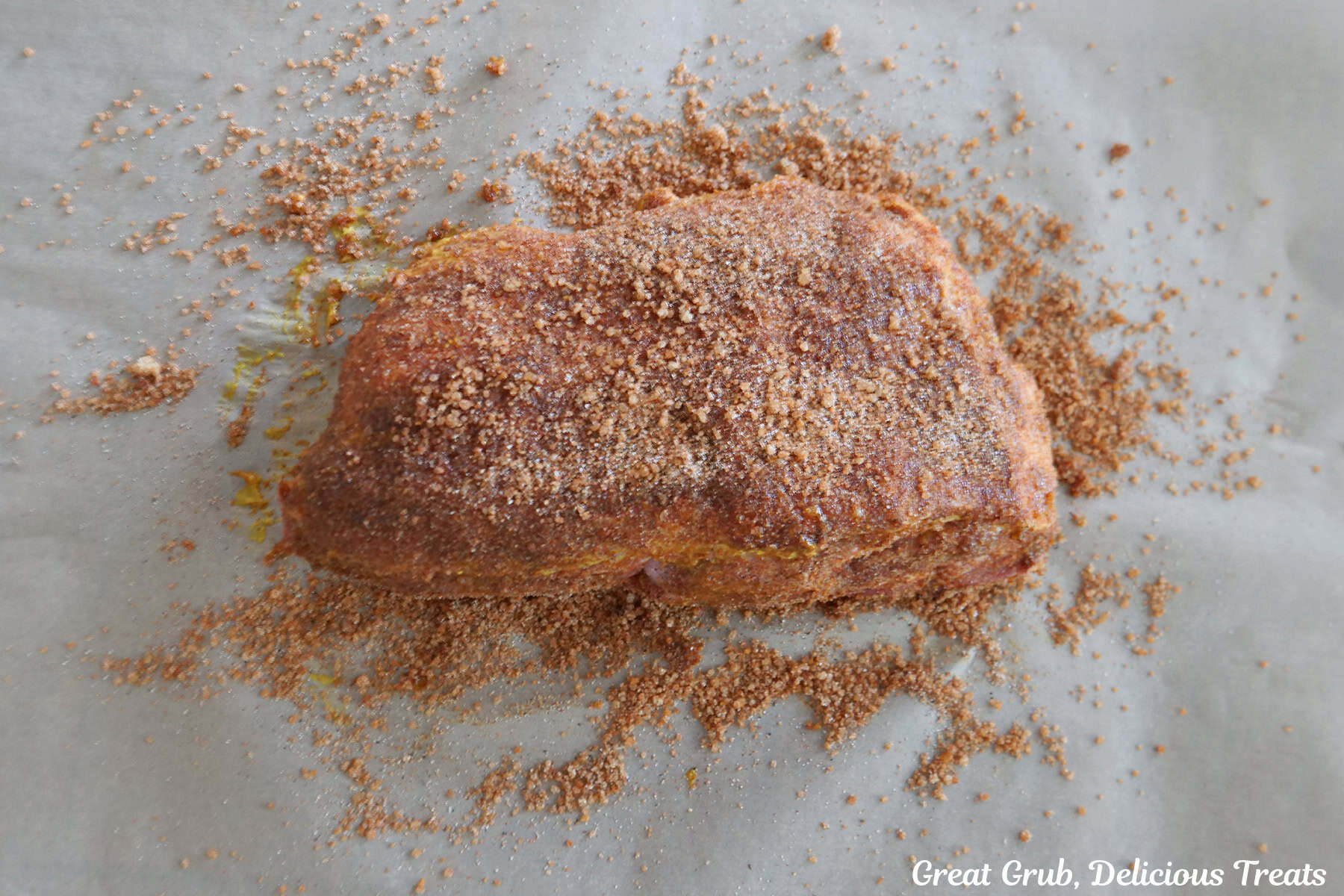 A pork roast placed on parchment paper that is covered in yellow mustard and a zesty blend of seasonings.