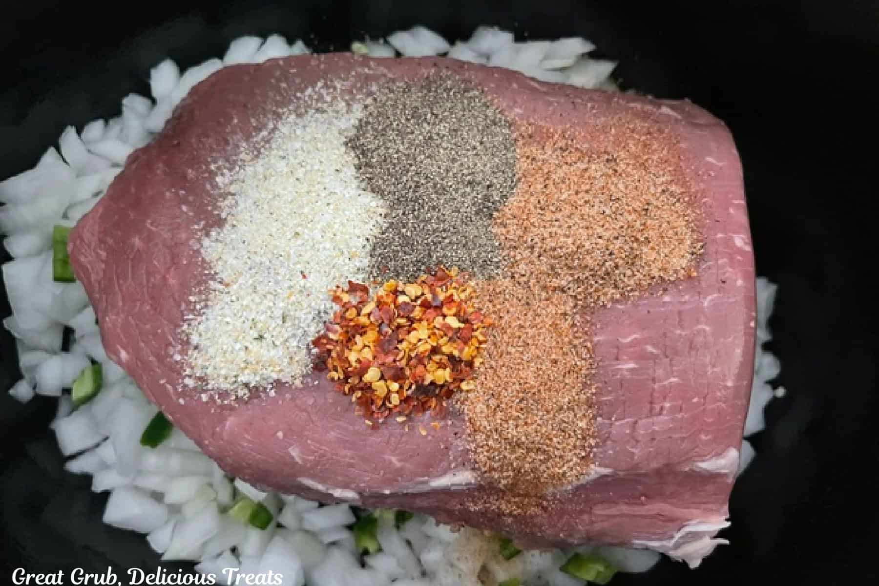 A crock pot filled with onions, jalapenos, a roast beef, and seasoning sprinkled on top of the roast.