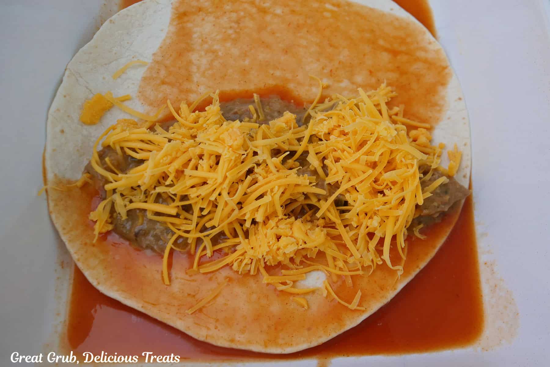 A white baking dish with a flour tortilla place in red sauce with shredded beef and shredded cheese on it.