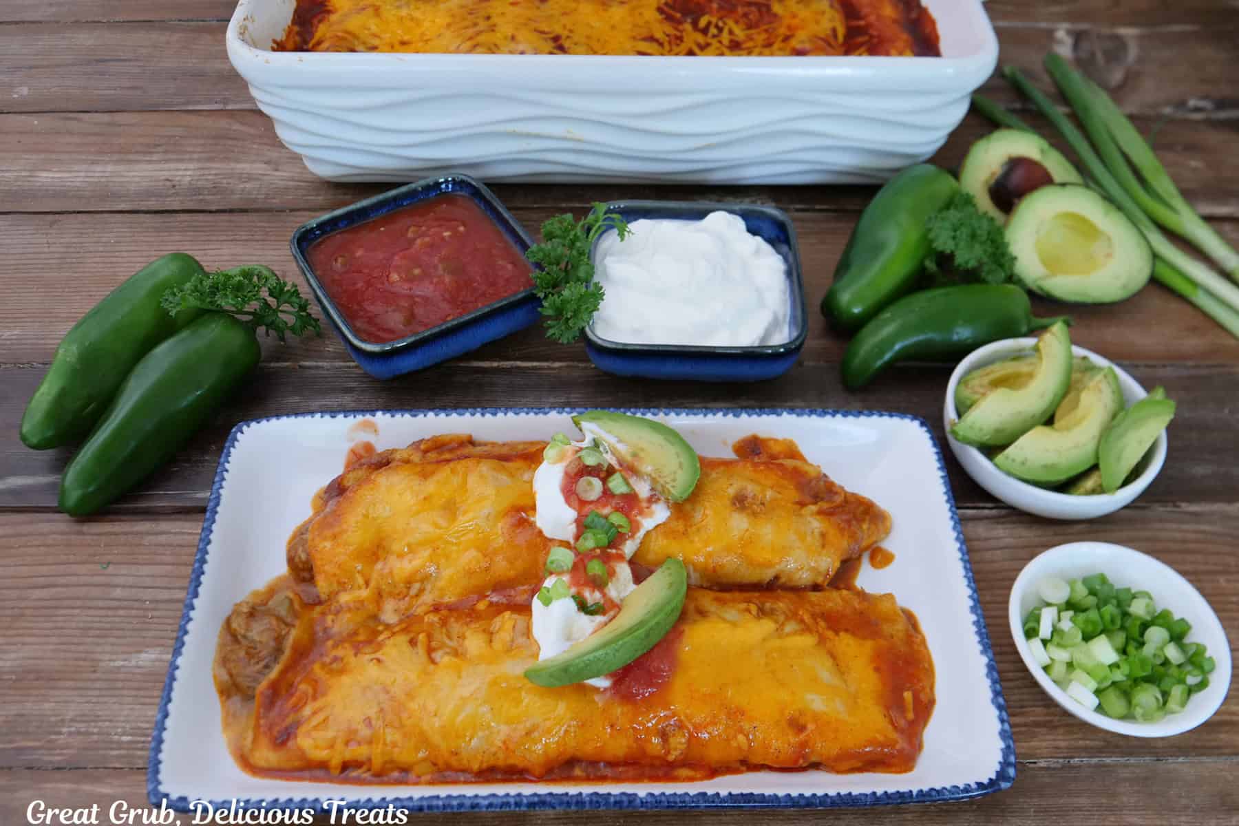 A wood surface with a white oblong plate with two enchiladas on it with a baking dish filled with enchiladas in the background and garnish ingredients placed randomly on the wood surface.