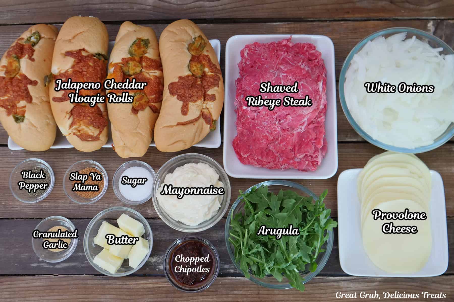 A rustic wood surface with all the ingredients placed on it to make steak sandwiches with caramelized onions and smoky aioli.