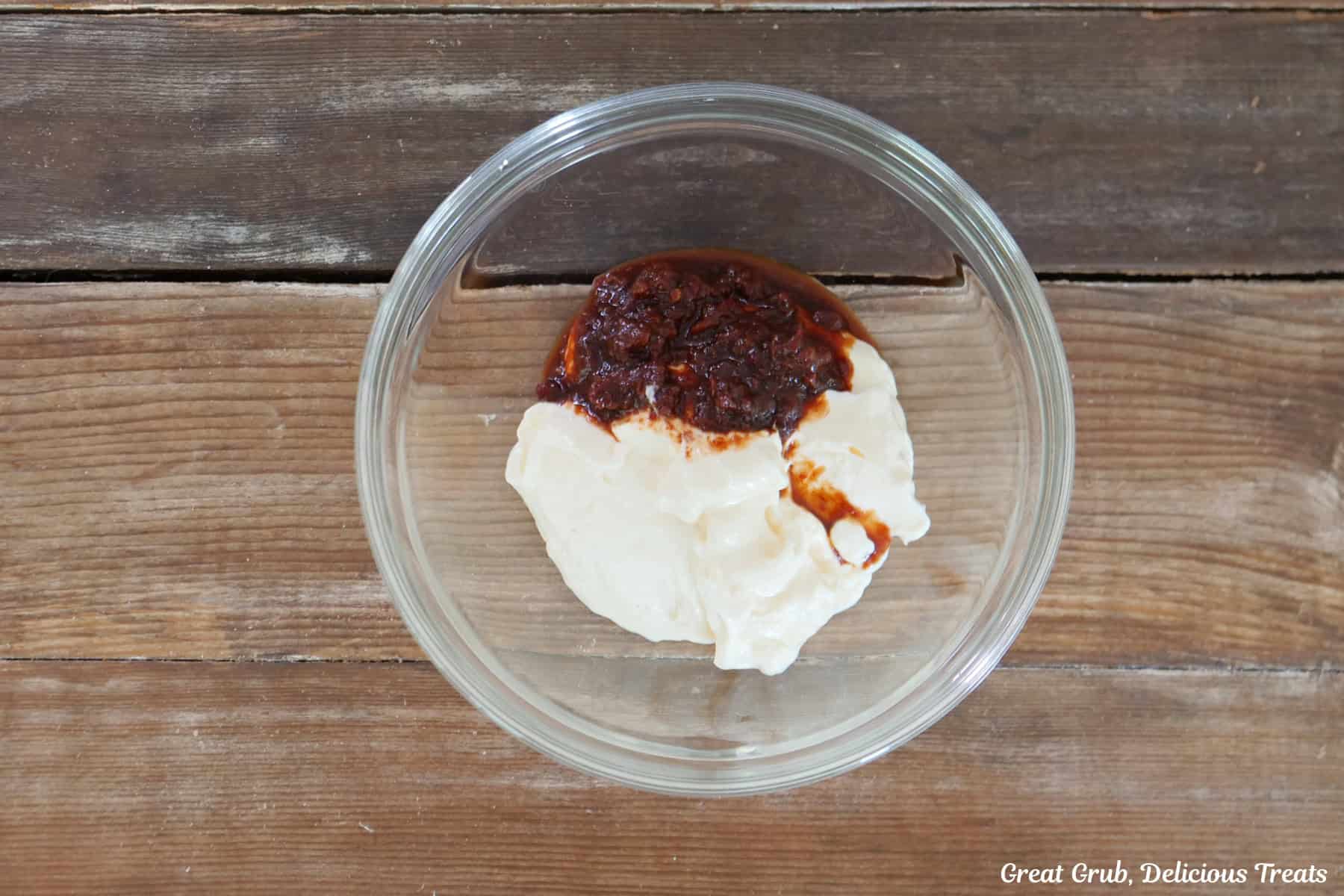 A wood surface with a glass bowl filled with mayonnaise, chipotles and their juice before being mixed together.