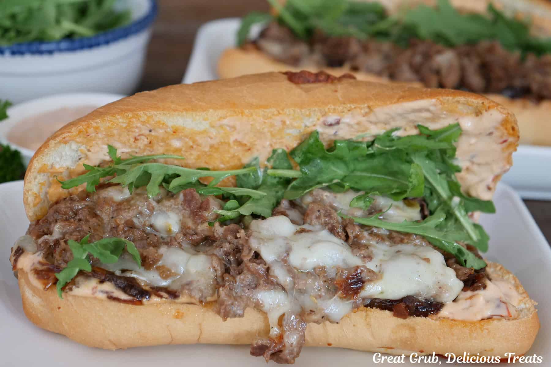 A hoagie roll cut in half stuffed with ribeye steak, cheese, arugula, and placed on a white plate.
