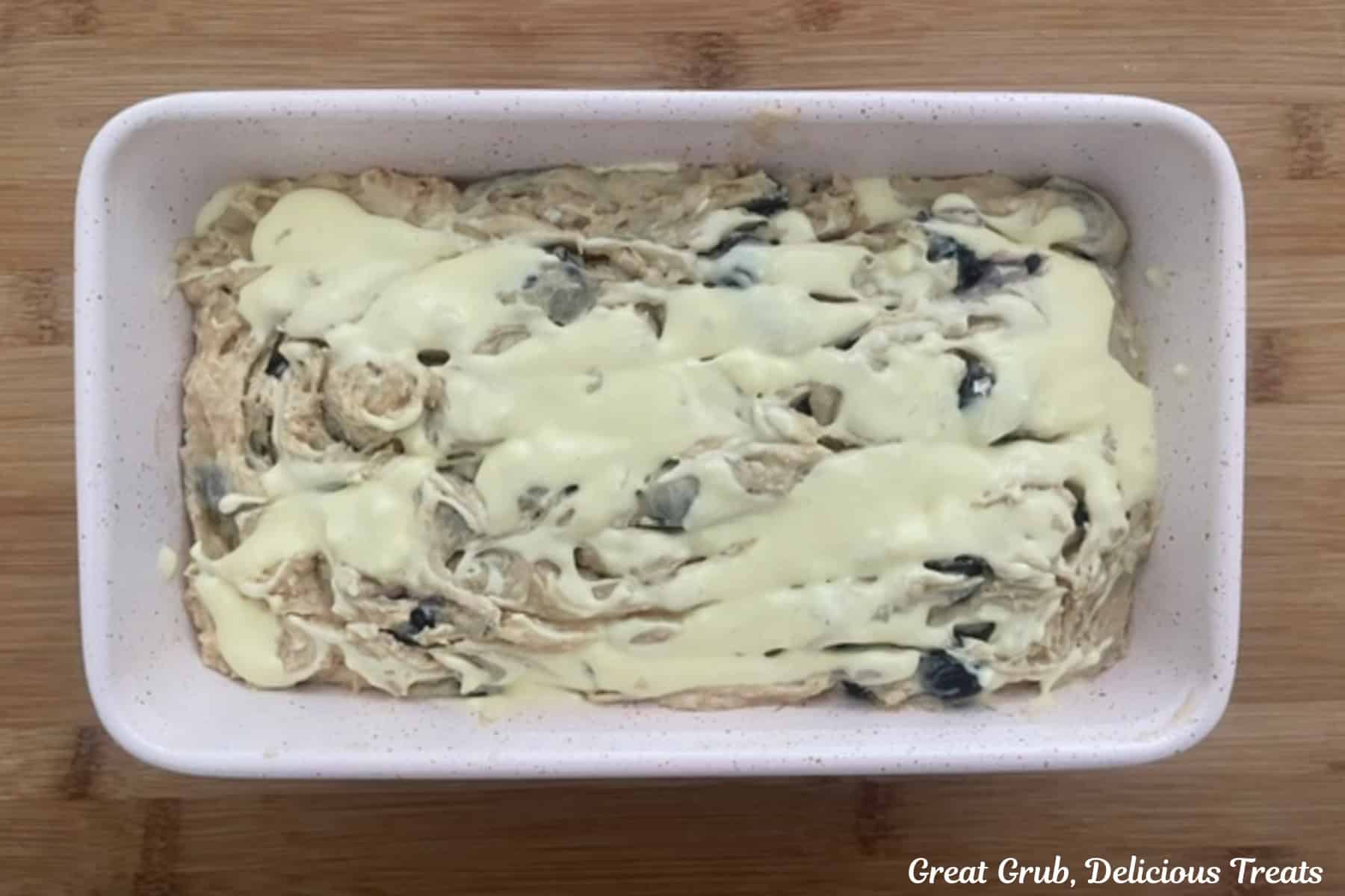 A wood surface with a 9 by 5 white baking loaf pan filled with blueberry cream cheese loaf before being baked in the oven.