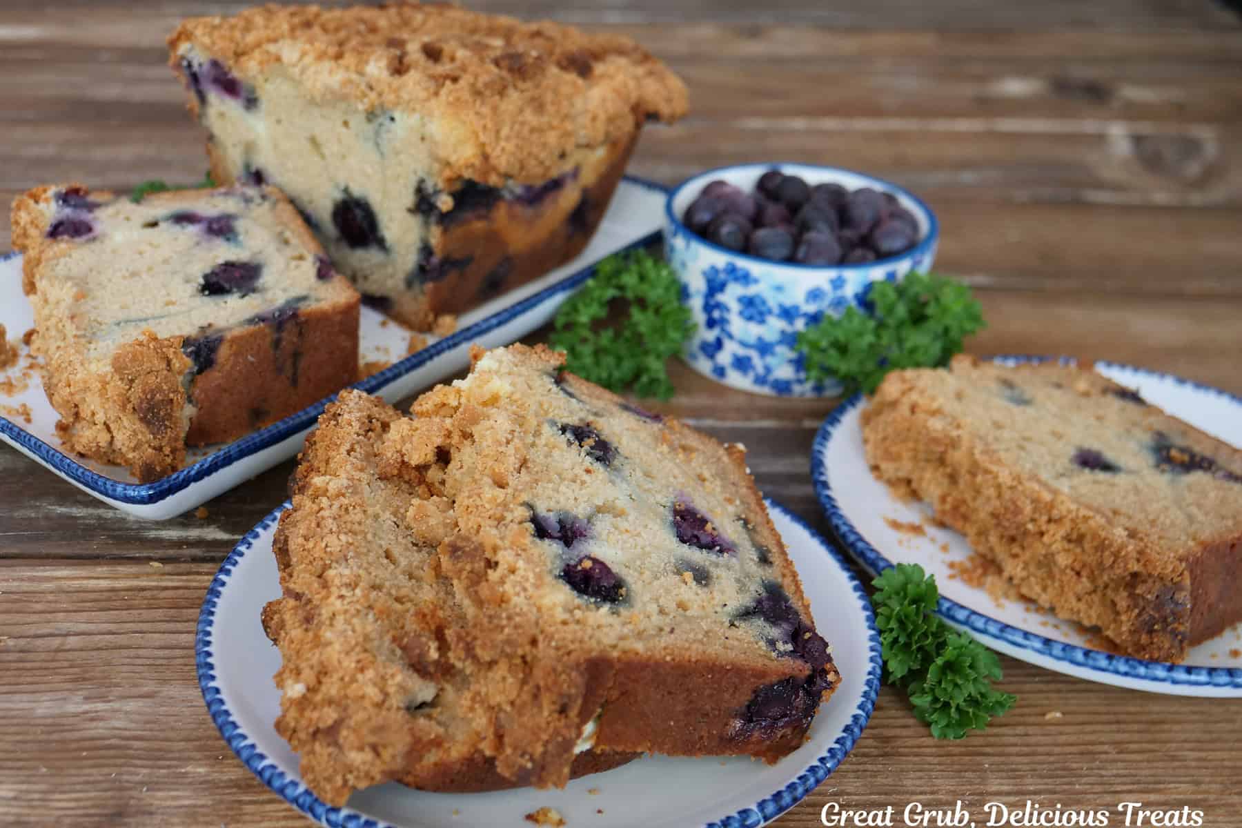 A wood surface with three white plates with slices of blueberry cream cheese loaf on it with a small white and blue floral bowl filled with blueberries.