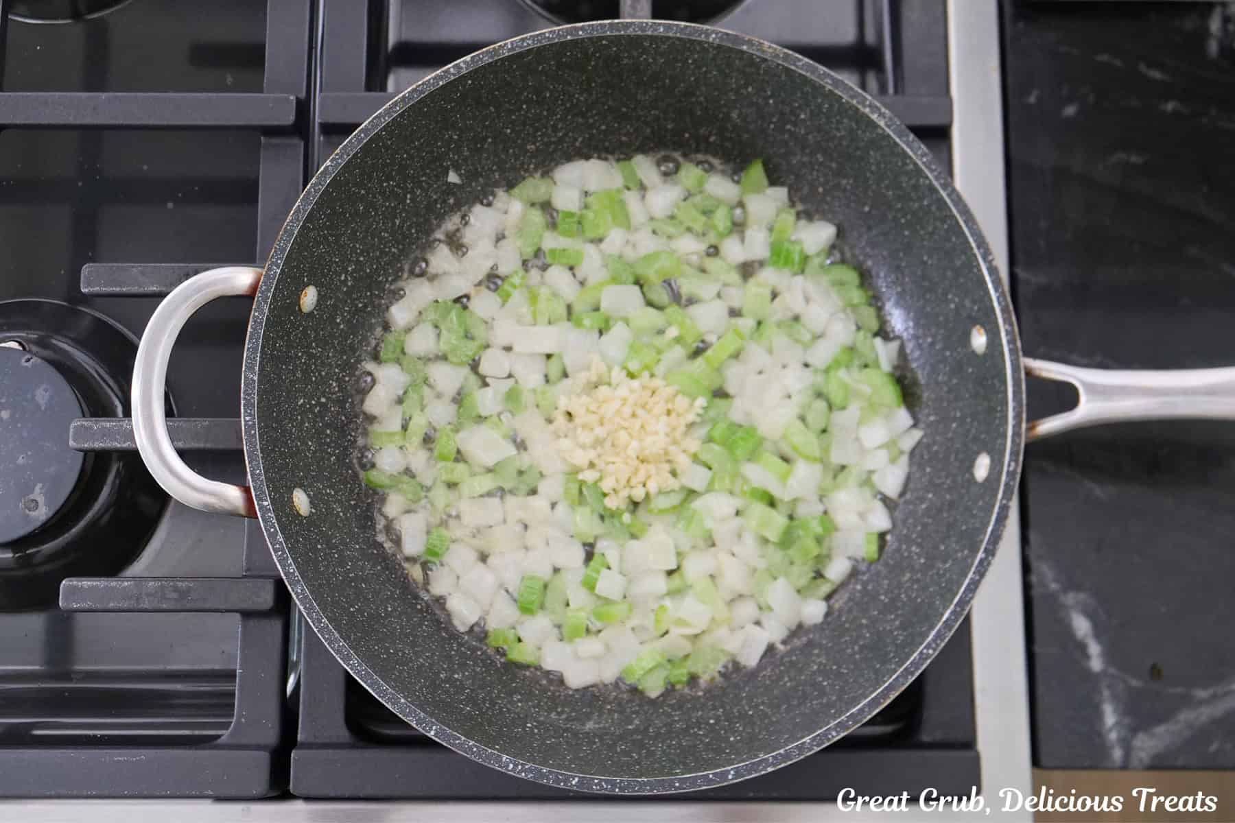 A frying pan on the stovetop with onions, celery, and garlic sauteing in it.