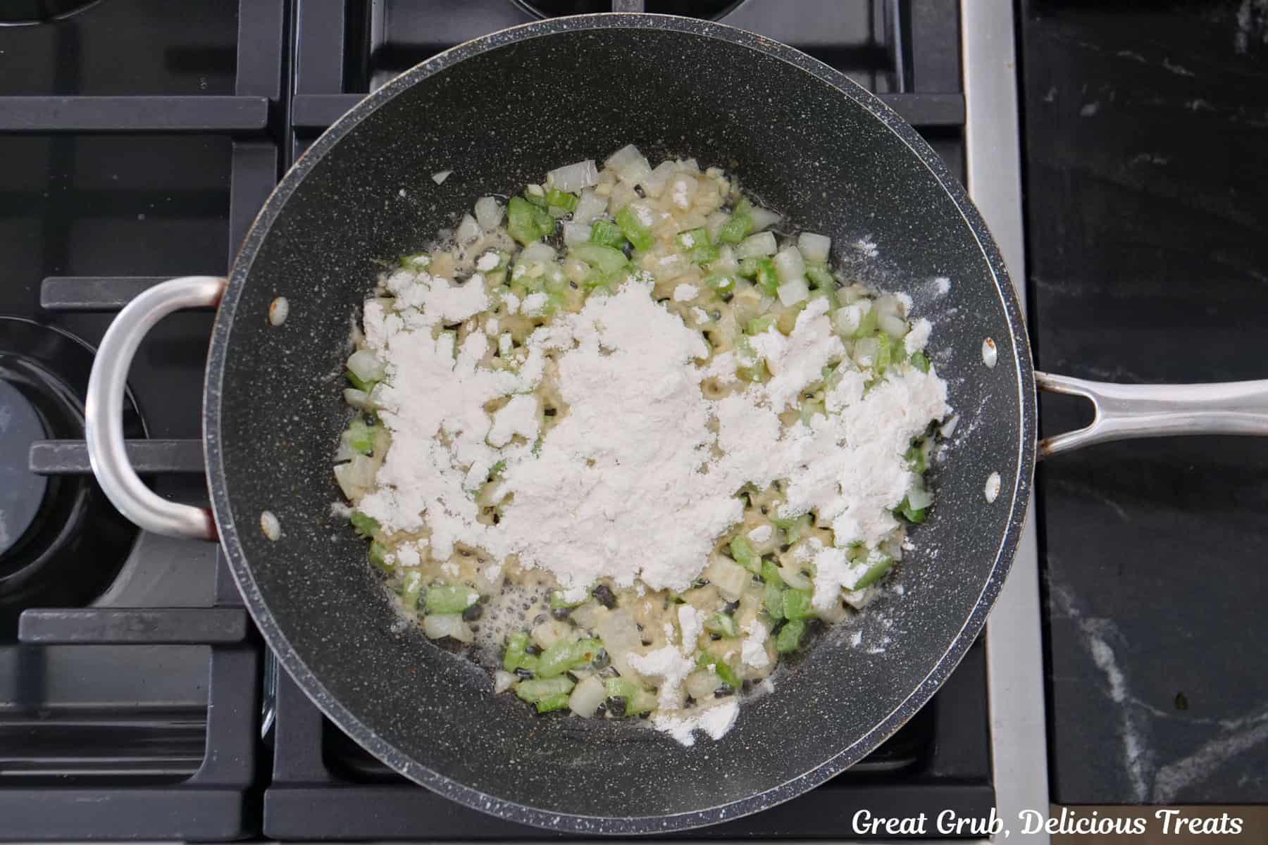 A frying pan on the stove with onions, celery, garlic, and flour sprinkled over the top.