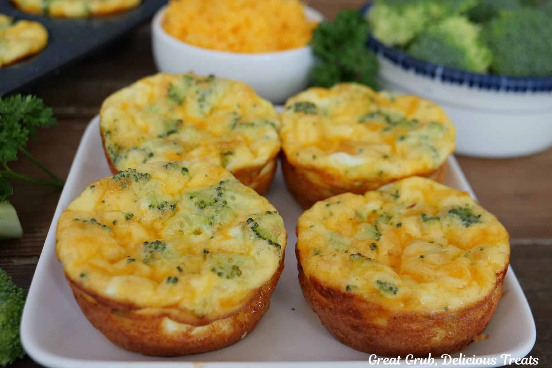 A small square white plate with four broccoli cheddar egg bites on it with broccoli and cheese in white bowls in the background.