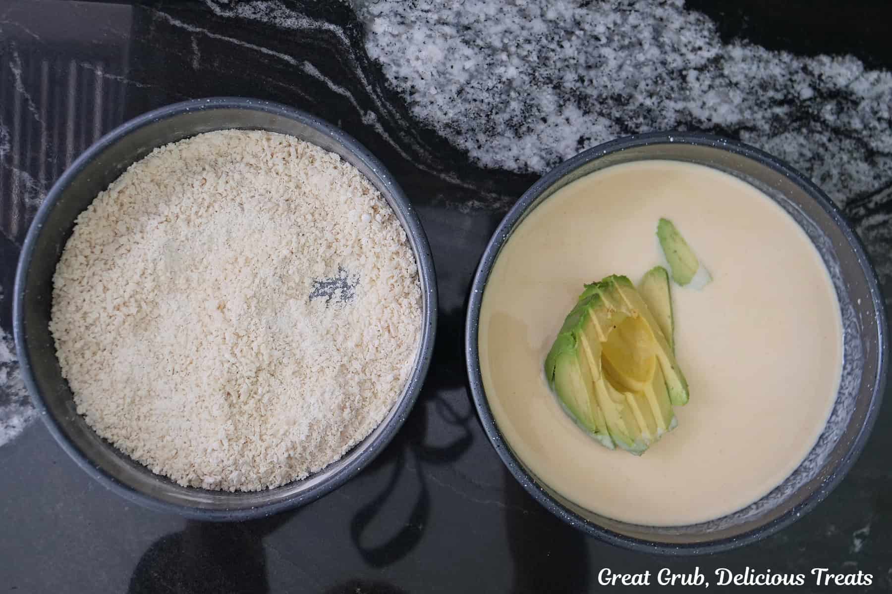 A black granite counter with two round pans one filled with seasoned breadcrumbs and the other with a buttermilk mixture with avocado slices in it.
