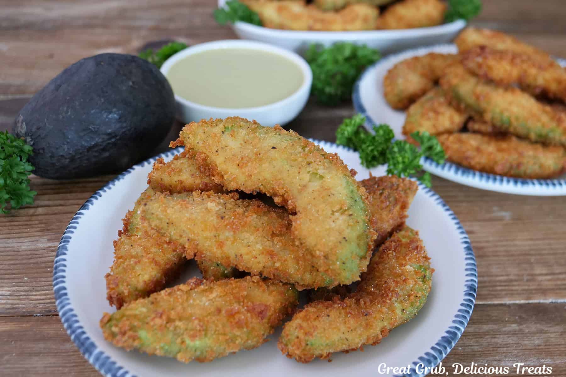 A wood surface with a few round white plates with blue trim with crispy avocado fries on them with a whole avocado and dipping sauce in the background.