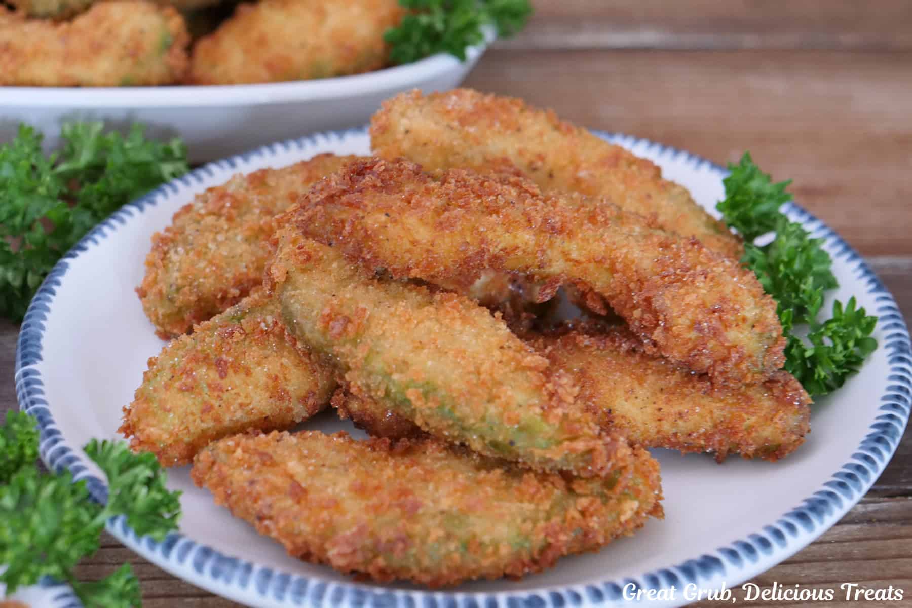 A wood surface with a round white plate with blue trim with about 8 crispy avocado fries on it.