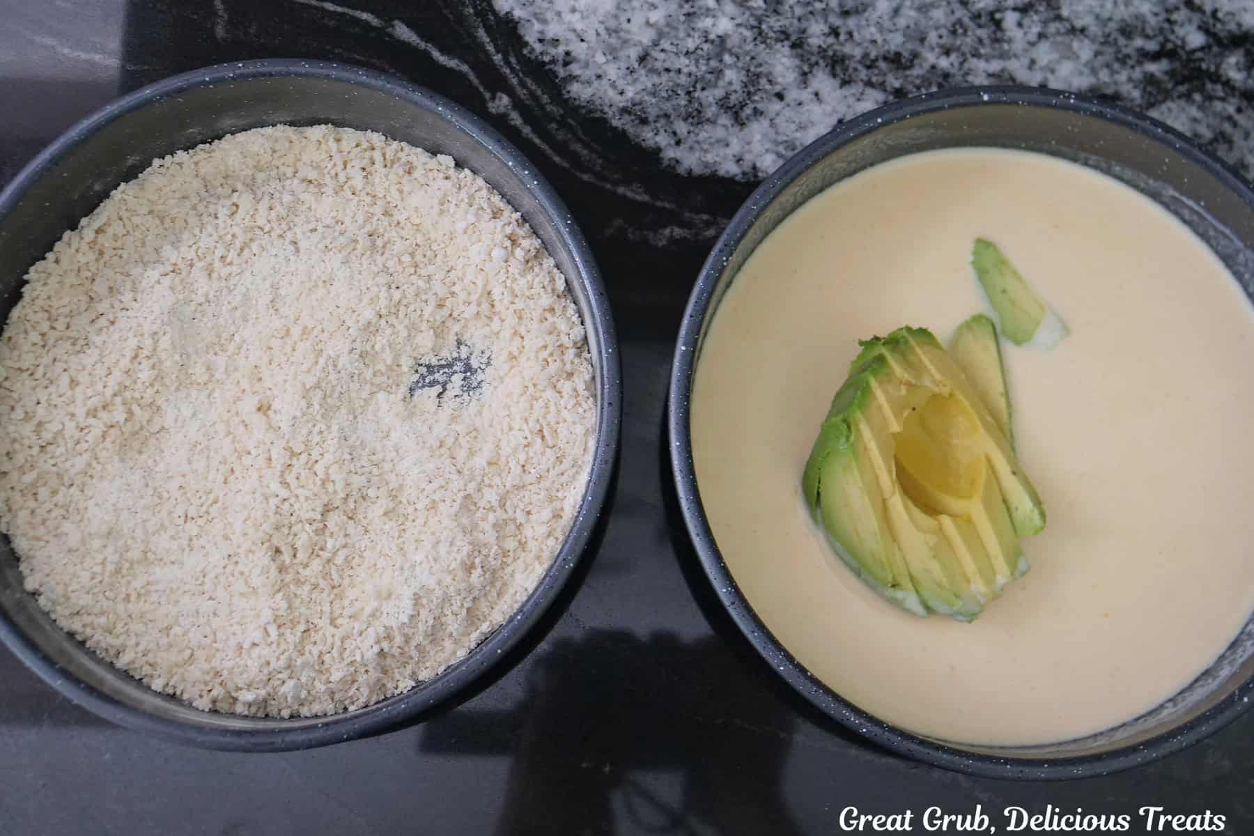 Two black metal round pans, one filled with a breadcrumb mixture and the other with a buttermilk mixture with an avocado in it.