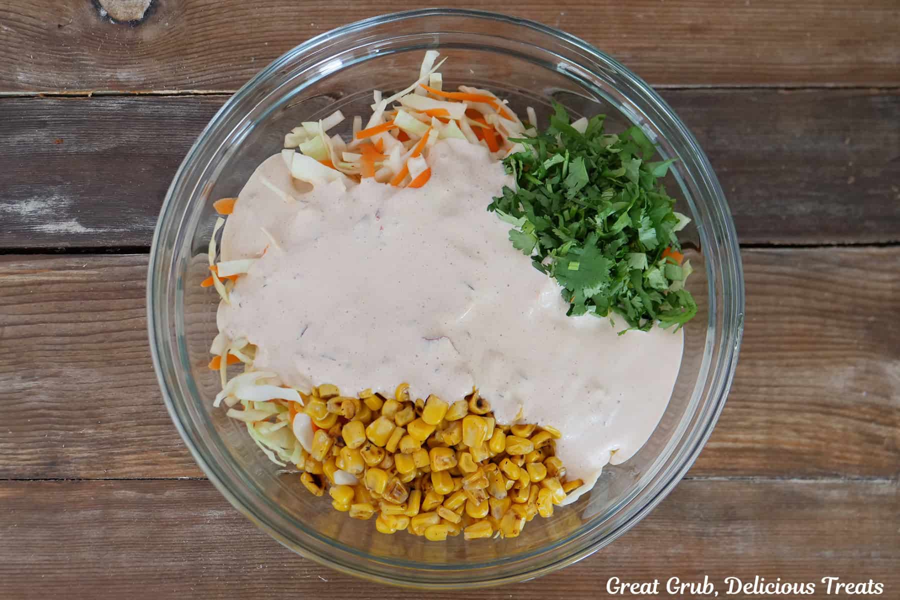 A wood surface with a glass bowl filled with corn, cilantro, coleslaw mix, shredded cheese, and a creamy sauce before it is all mixed together.