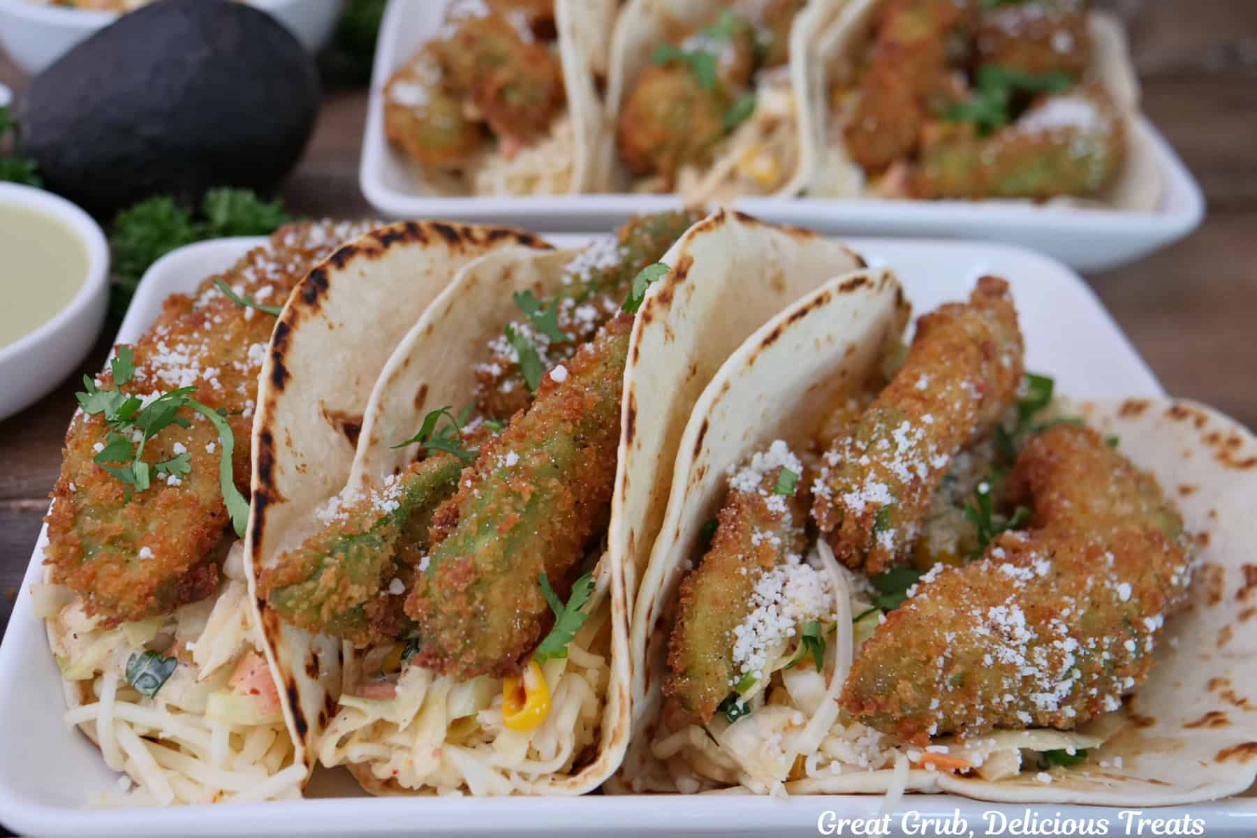 Two oblong white plates with three crispy avocado tacos on each plate.