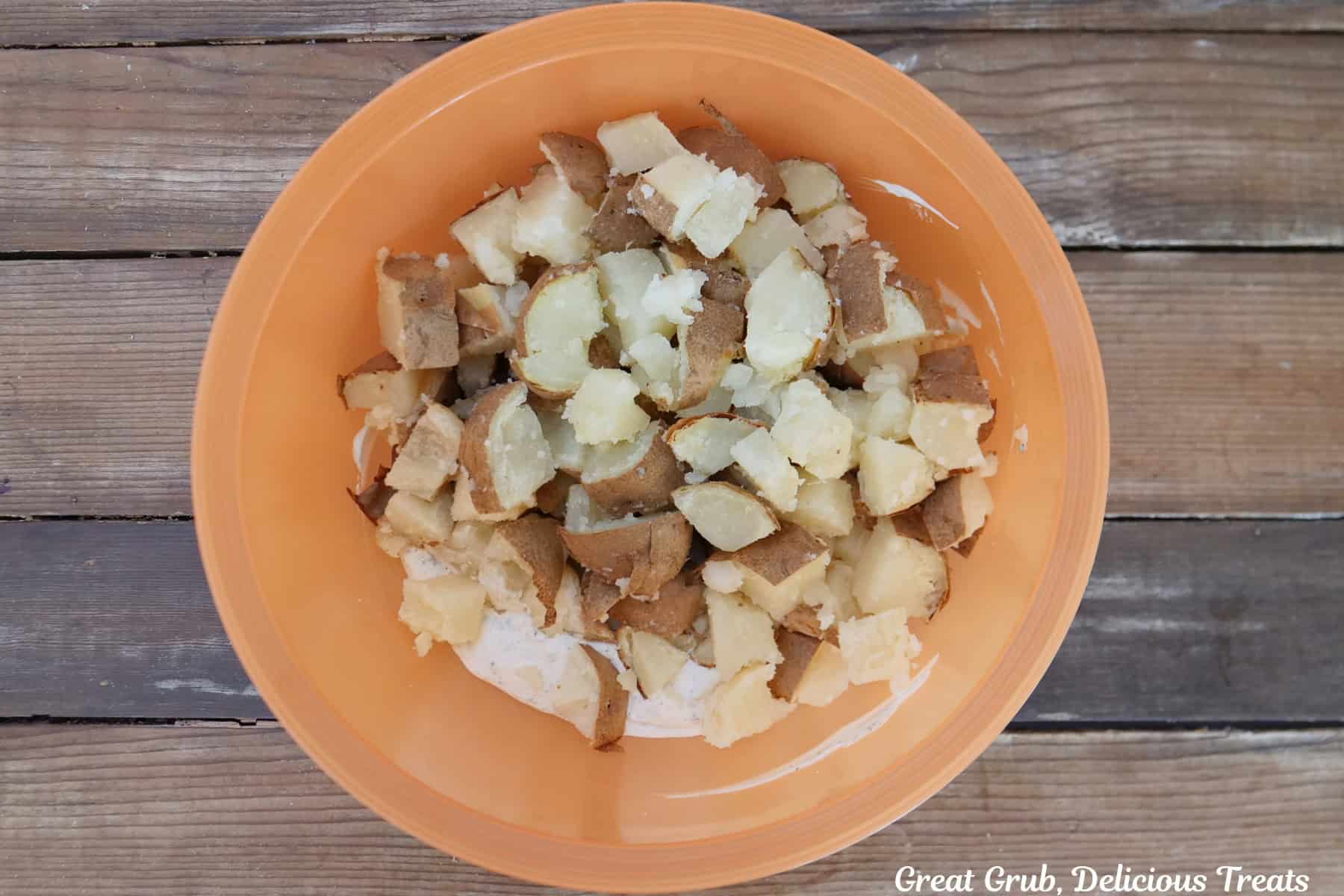 A wood surface with an orange mixing bowl filled with diced potatoes and the creamy sauce before being combined.