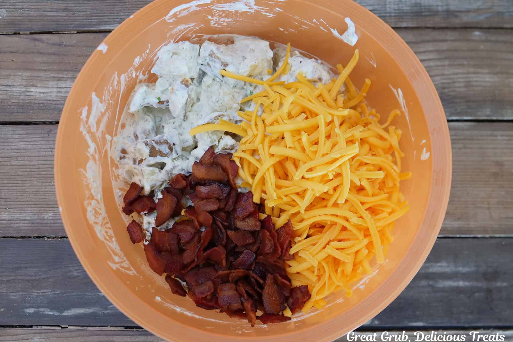 A wood surface with an orange bowl will the shredded cheddar and crispy bacon added to the potato salad mixture.