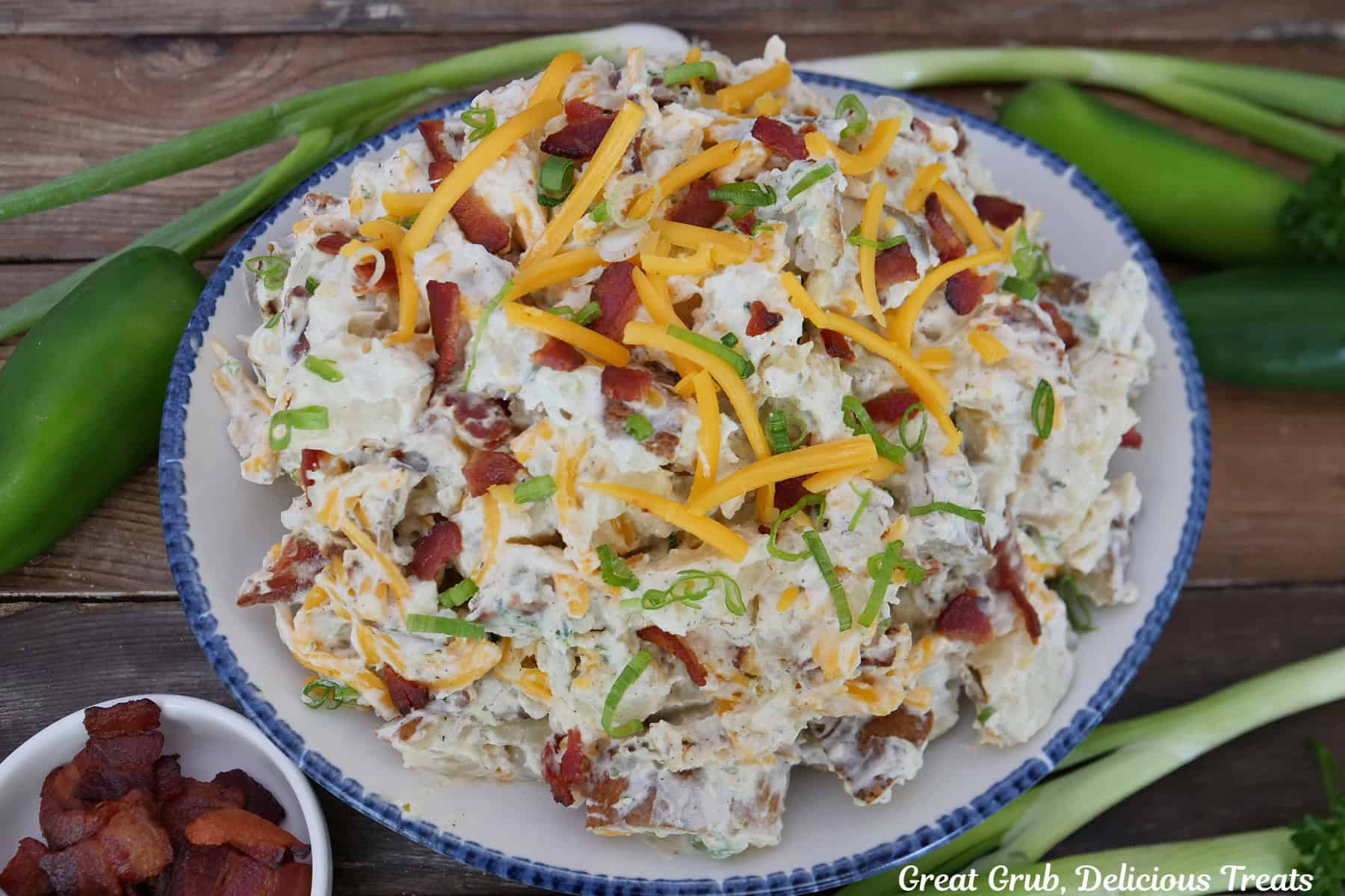 A blue trimmed white bowl with a serving of loaded baked potato salad in it and placed on a rustic wood surface.