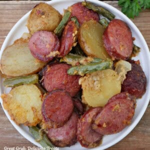 A white small round plate with a serving of Parmesan crusted smoked sausage, potatoes, and green beans on it.