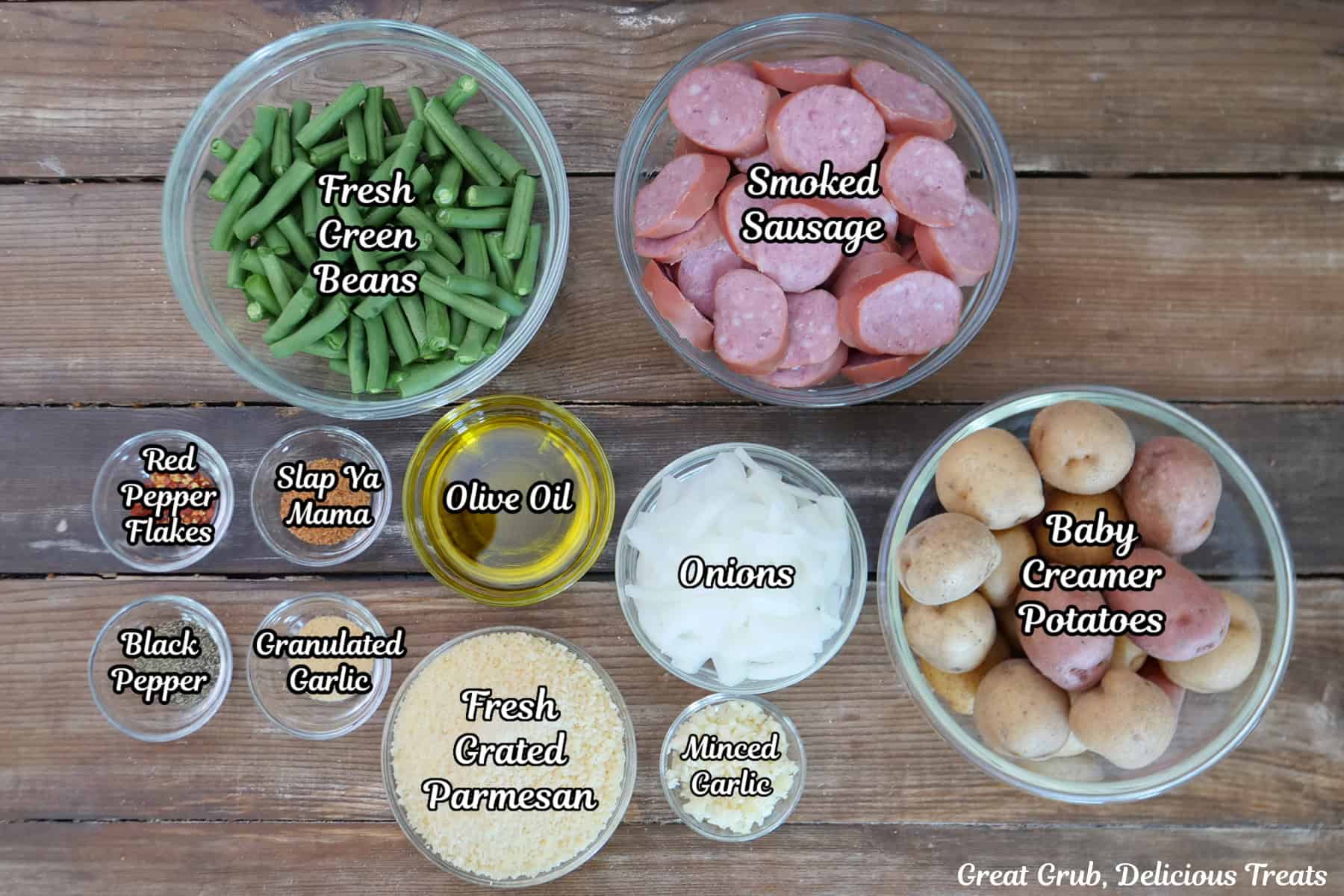 A rustic wood surface with all the ingredients placed on it to make sheet pan Parmesan crusted smoked sausage and potatoes.