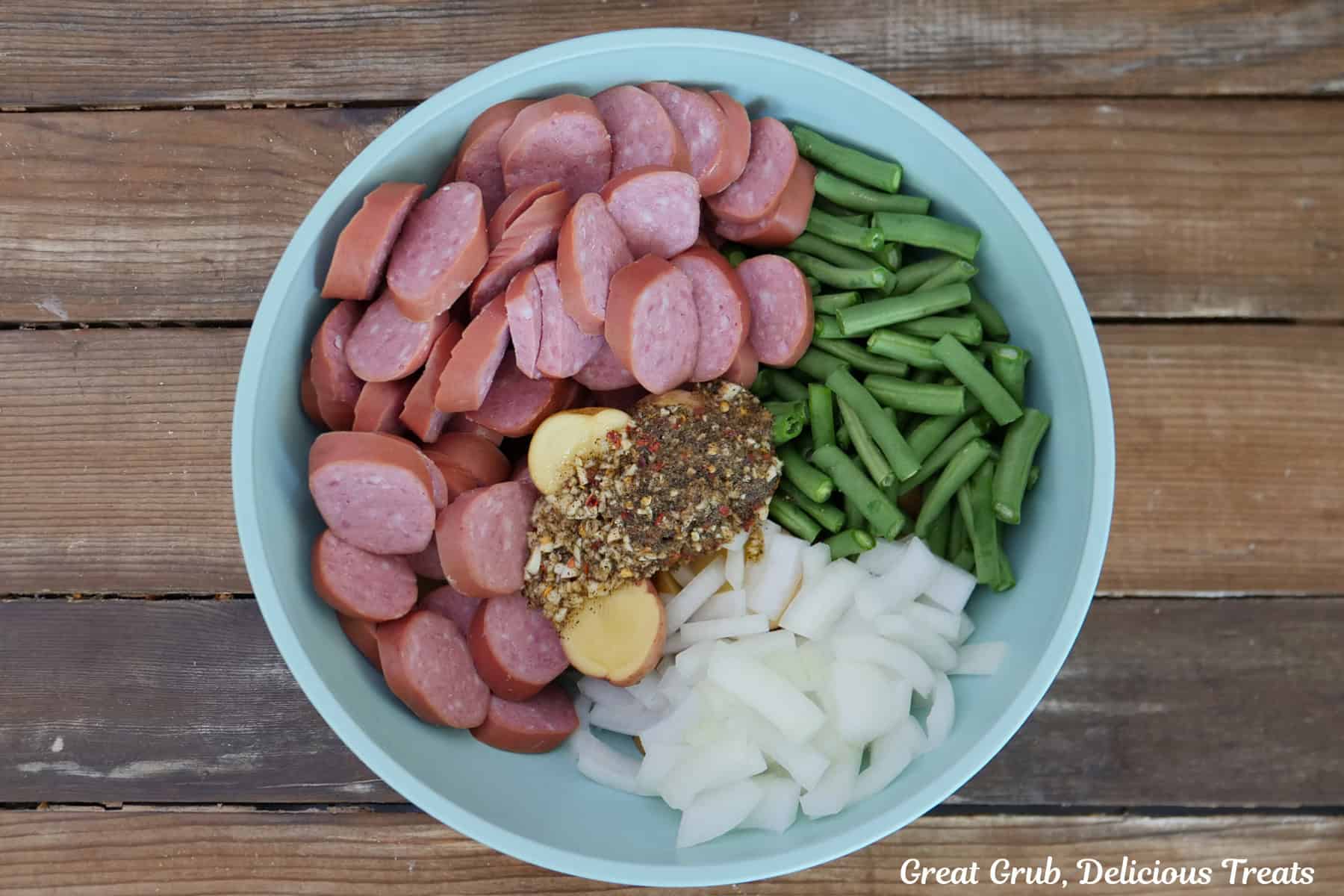 A wood surface with a light blue bowl filled with sliced smoked sausage, potatoes, green beans, onions, and seasonings.