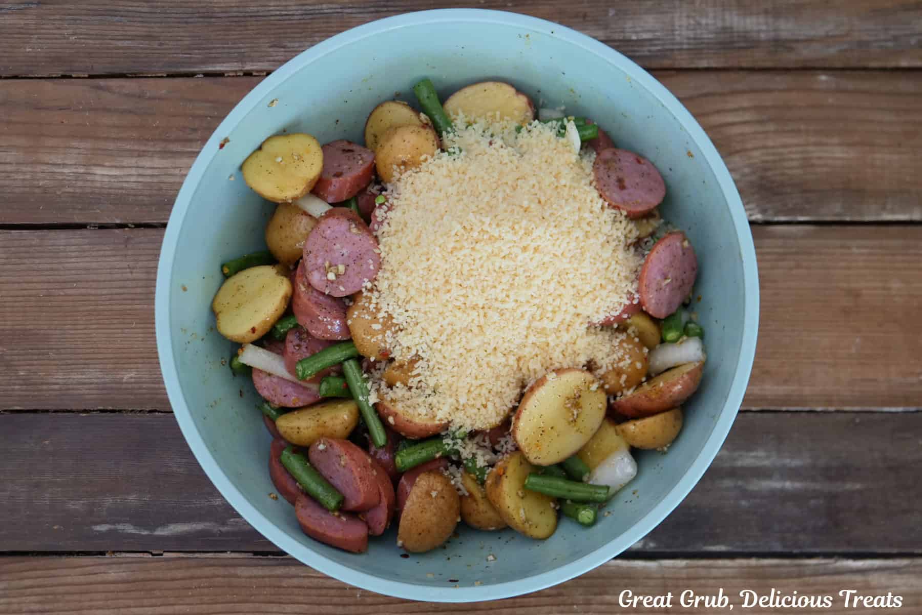 A wood surface with a light blue bowl will all the ingredients placed in it to make Parmesan crusted smoked sausage and potatoes.