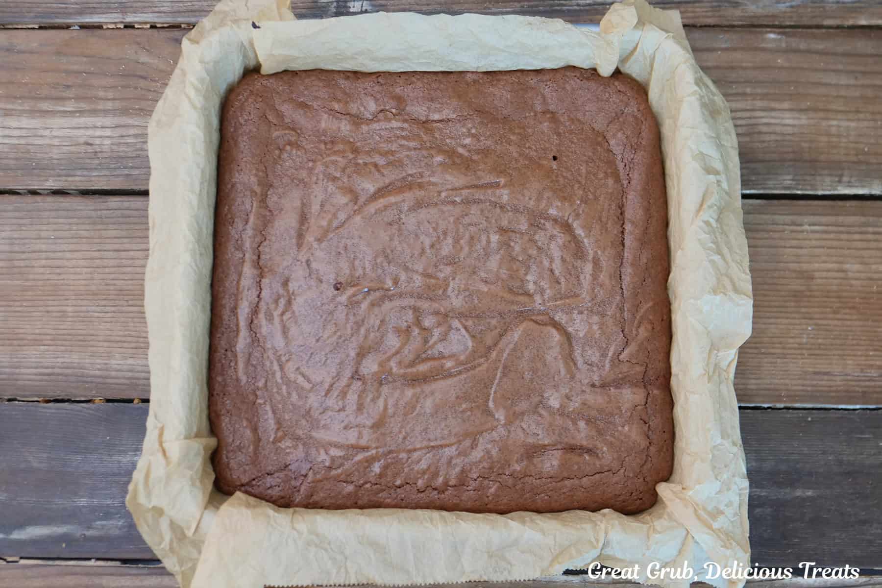 A rustic wood surface with a 9 by 9 baking dish on it, lined with parchment paper, with the brownie batter in it.