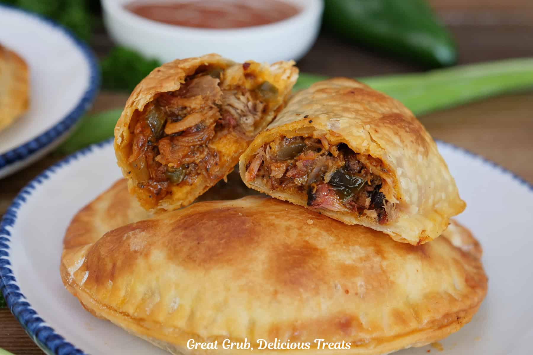 A round white plate with blue trim with three pulled pork empanadas on it with the one on top broke in half showing the inside ingredients.