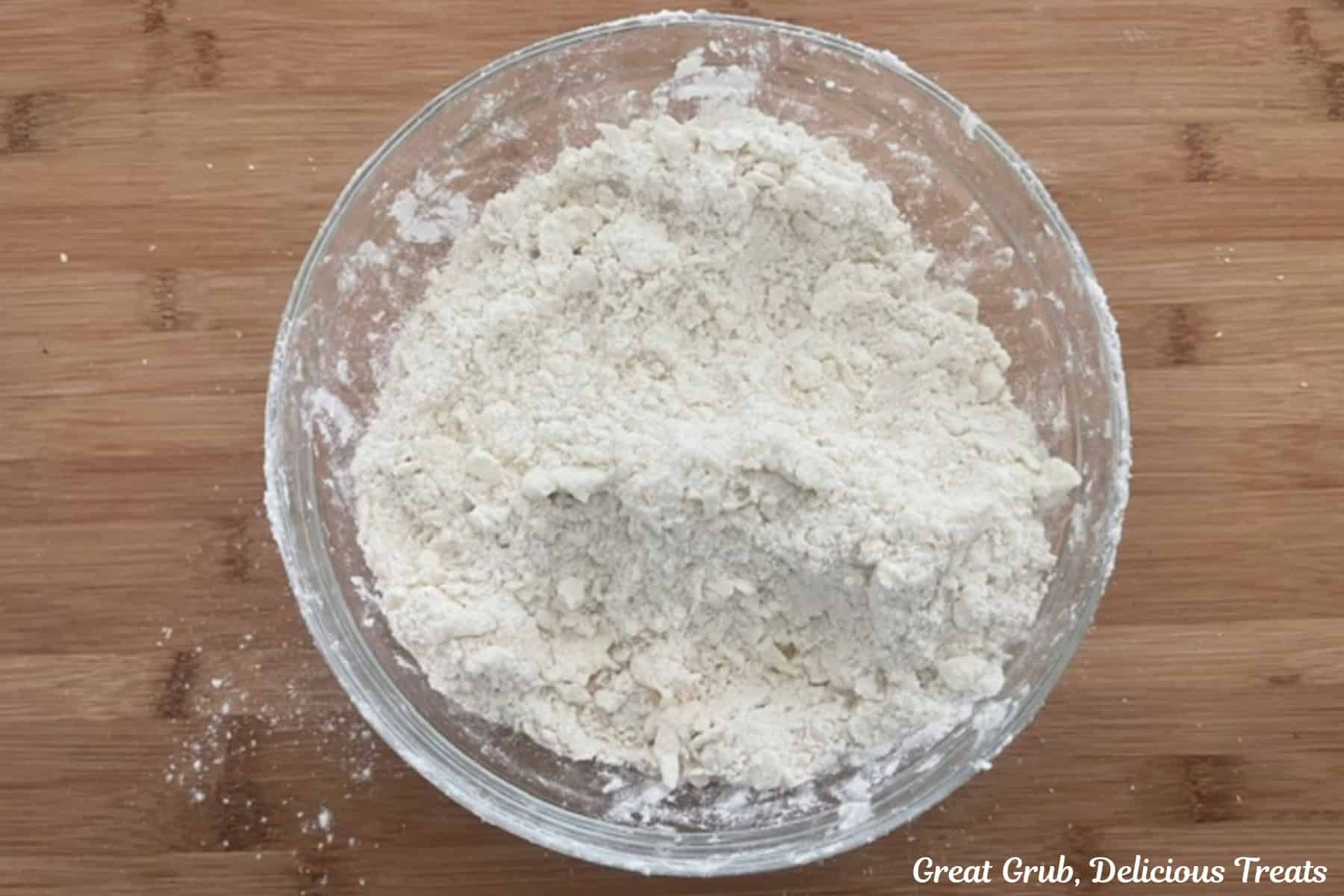 A wood cutting board with a glass bowl filled with flour mixture and butter combined into coarse crumbs.