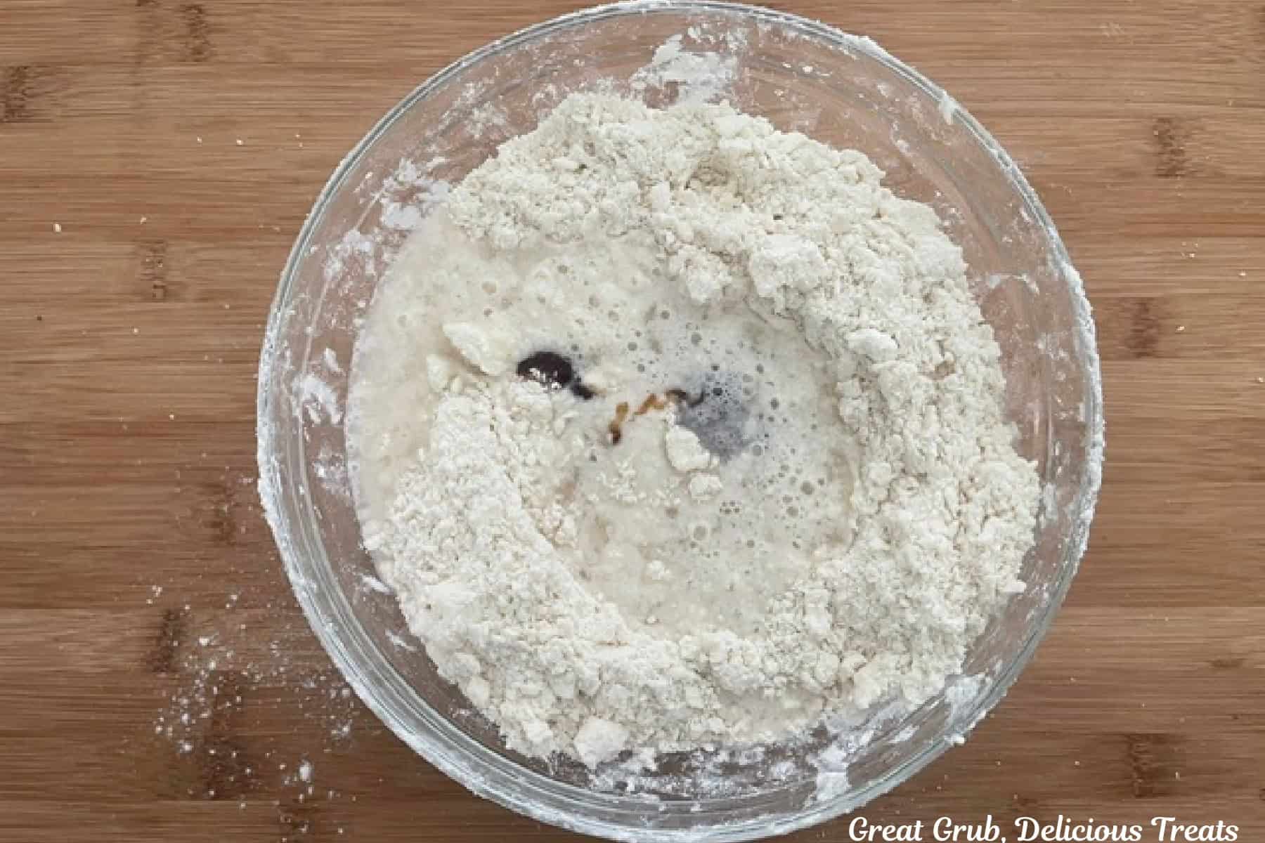 A wood cutting board with a glass bowl with flour mixture and vanilla bean paste added to it.
