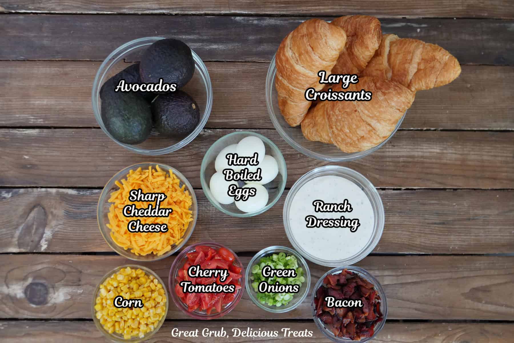 A wood surface with all the ingredients placed on it to make avocado salad croissant sandwiches.