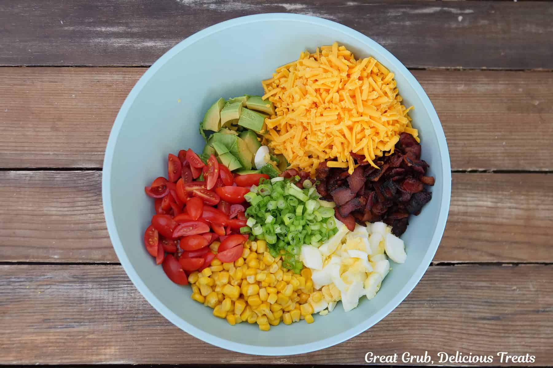 A wood surface with a light blue bowl filled with shredded cheese, bacon, avocado, corn, tomatoes, hardboiled eggs.