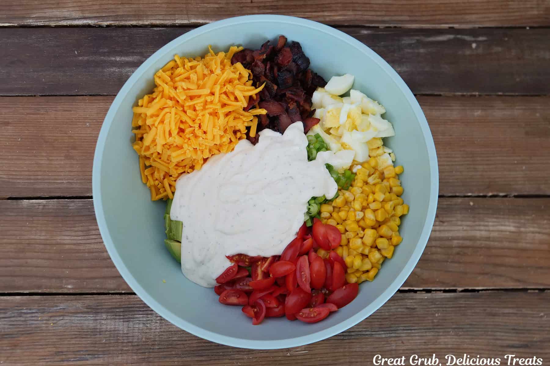 A wood surface with a light blue bowl filled with shredded cheese, bacon, avocado, corn, tomatoes, hardboiled eggs and ranch dressing.